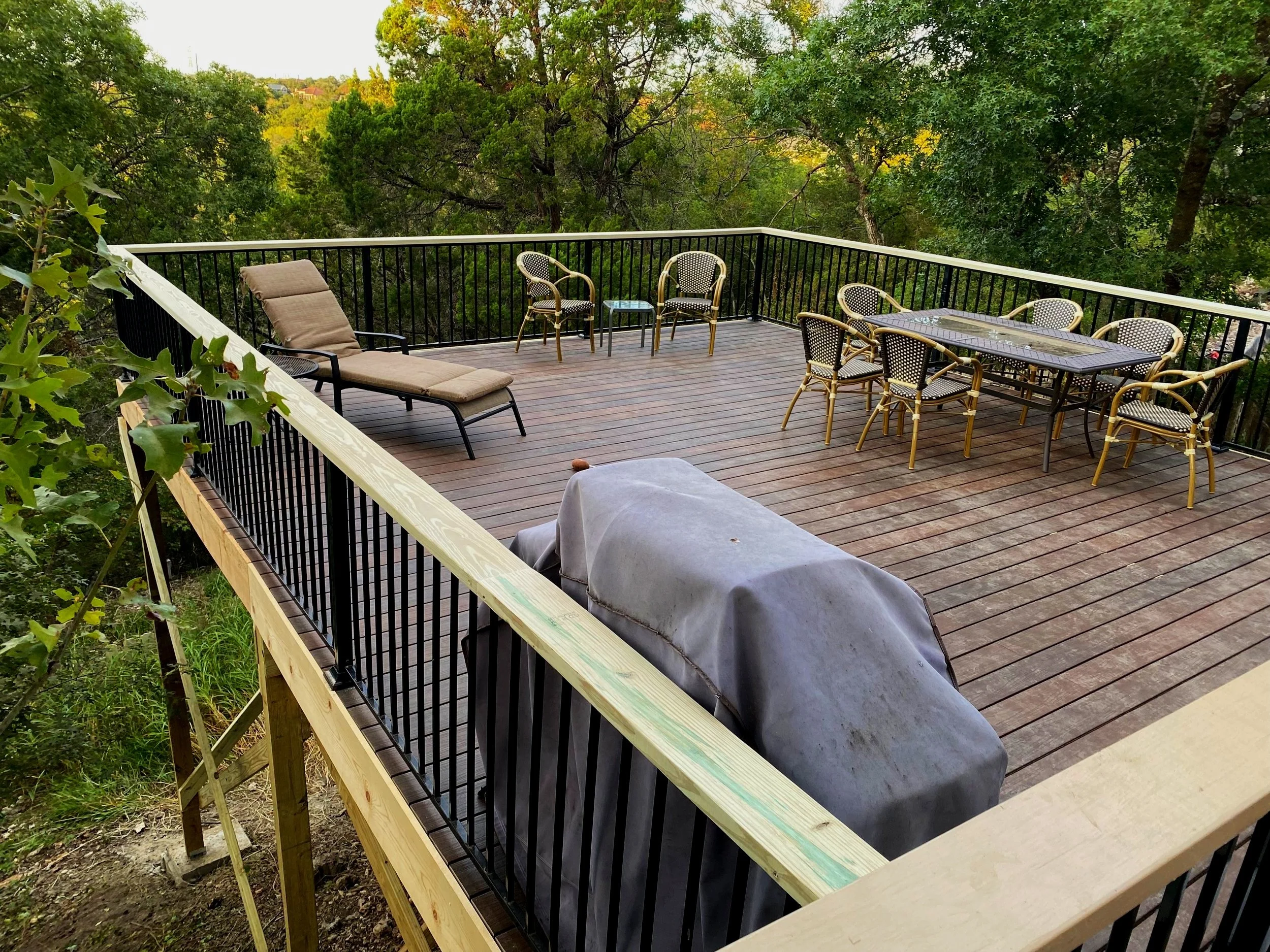A spacious outdoor wooden deck with black metal railing, overlooking a green forested area. The deck has outdoor furniture including a beige lounge chair with cushions, a large rectangular dining table with six matching chairs, and a covered grill.