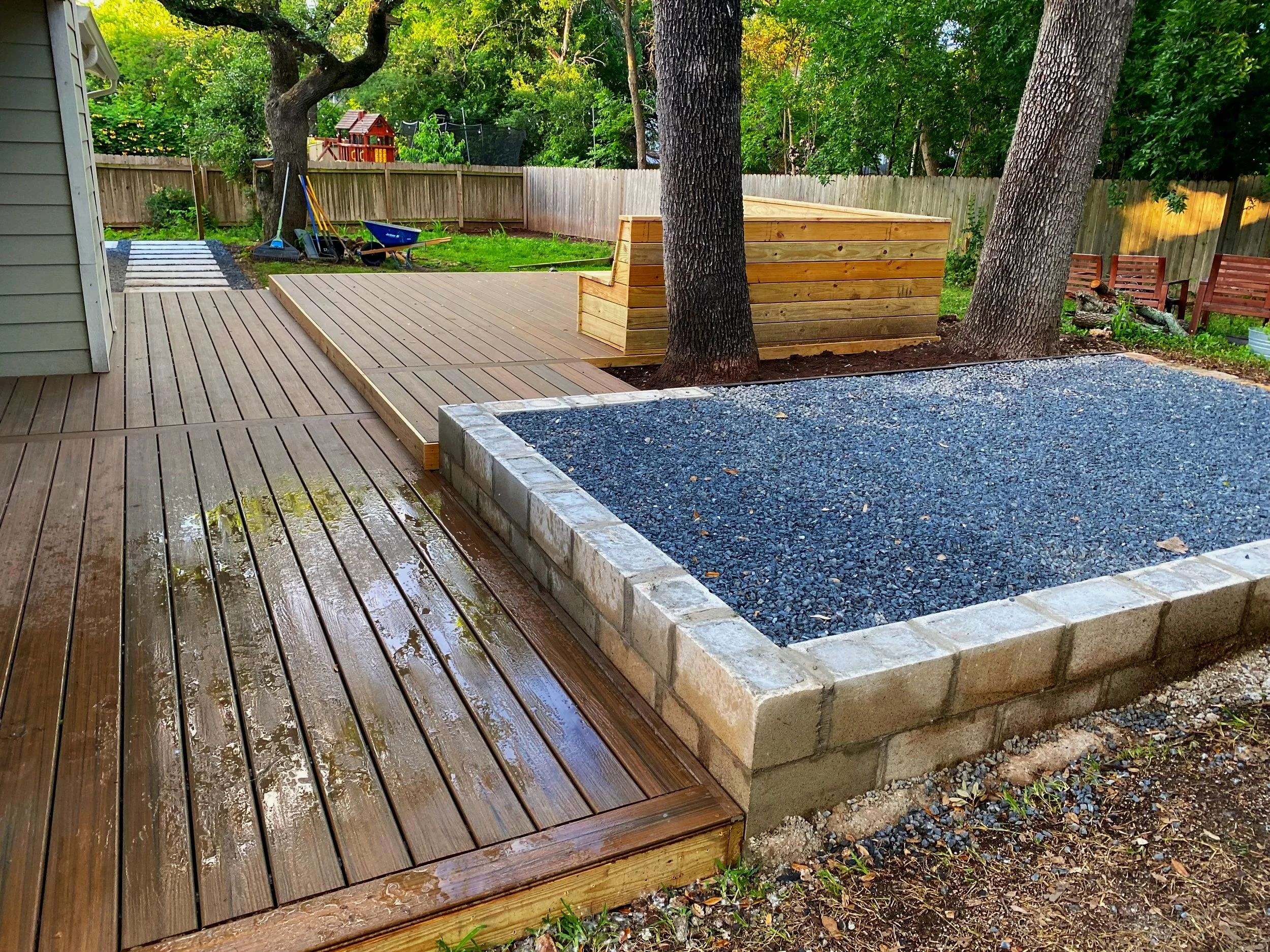 Composite ground level deck with a built-in cedar bench dining area and wrap-around stairs. A raised gravel pavilion was built beside the deck to accommodate a fire-pit and grill. A walkway leading off the property features custom concrete pavers.