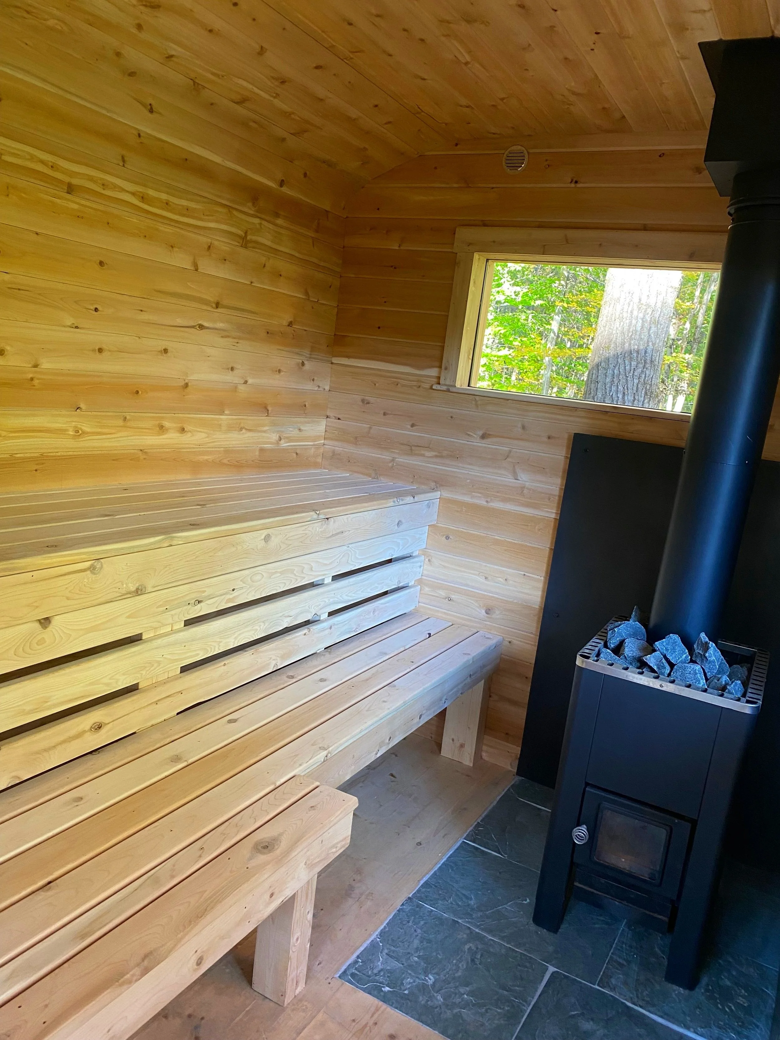 Interior of a small wooden sauna room with a wooden bench and a black wood stove with rocks, window showing green trees outside.