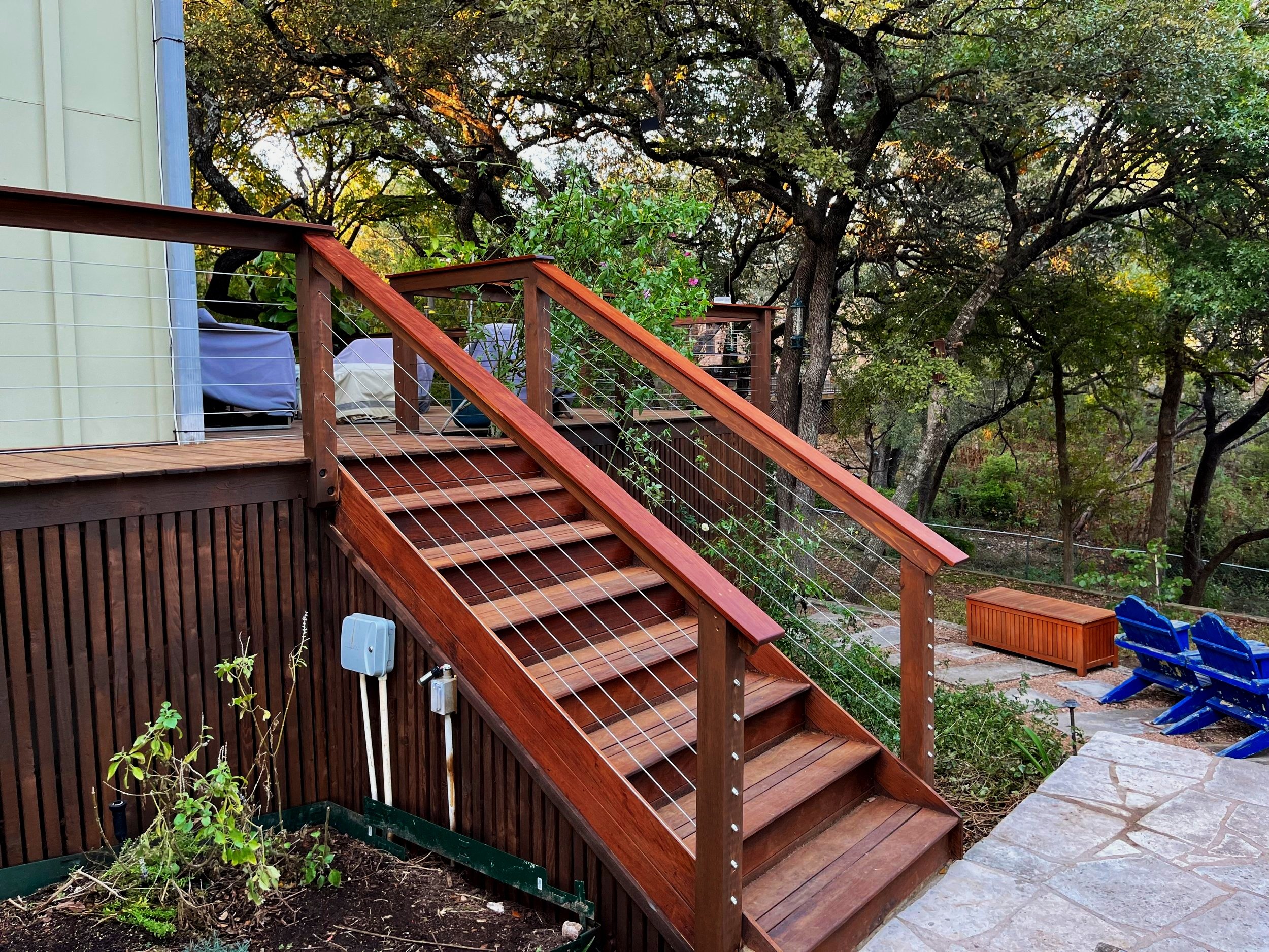 Wooden outdoor staircase with cable railing leading down to a landscaped patio area with blue chairs, a wooden bench, and surrounding trees.