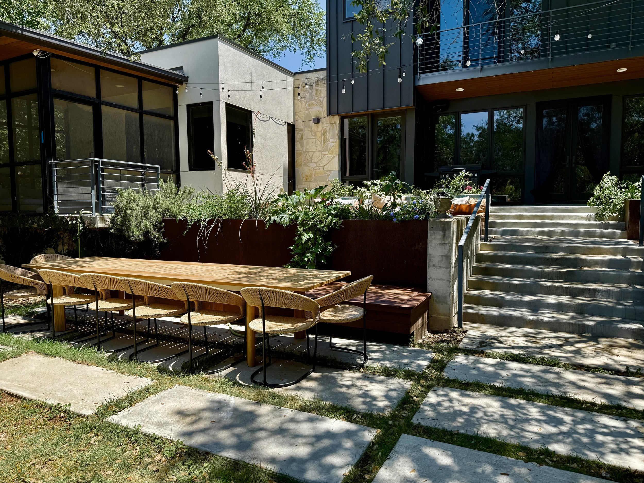 Outdoor patio with a long wooden table and eight matching chairs, surrounded by greenery and modern house with large glass windows and outdoor stairs.