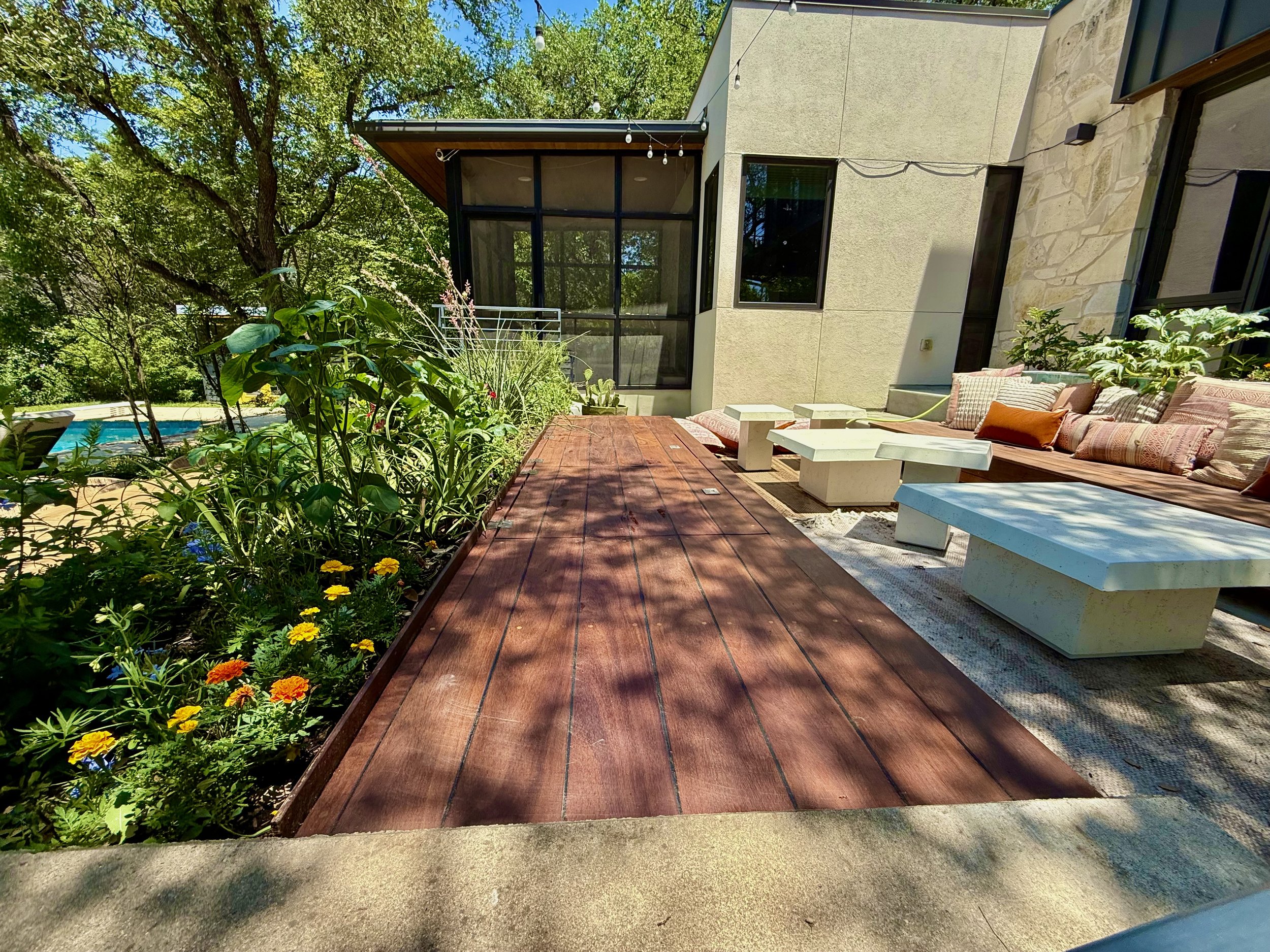 A backyard patio with a long wooden table, built-in white concrete benches, colorful cushions, and lush green plants, with a pool and trees in the background.