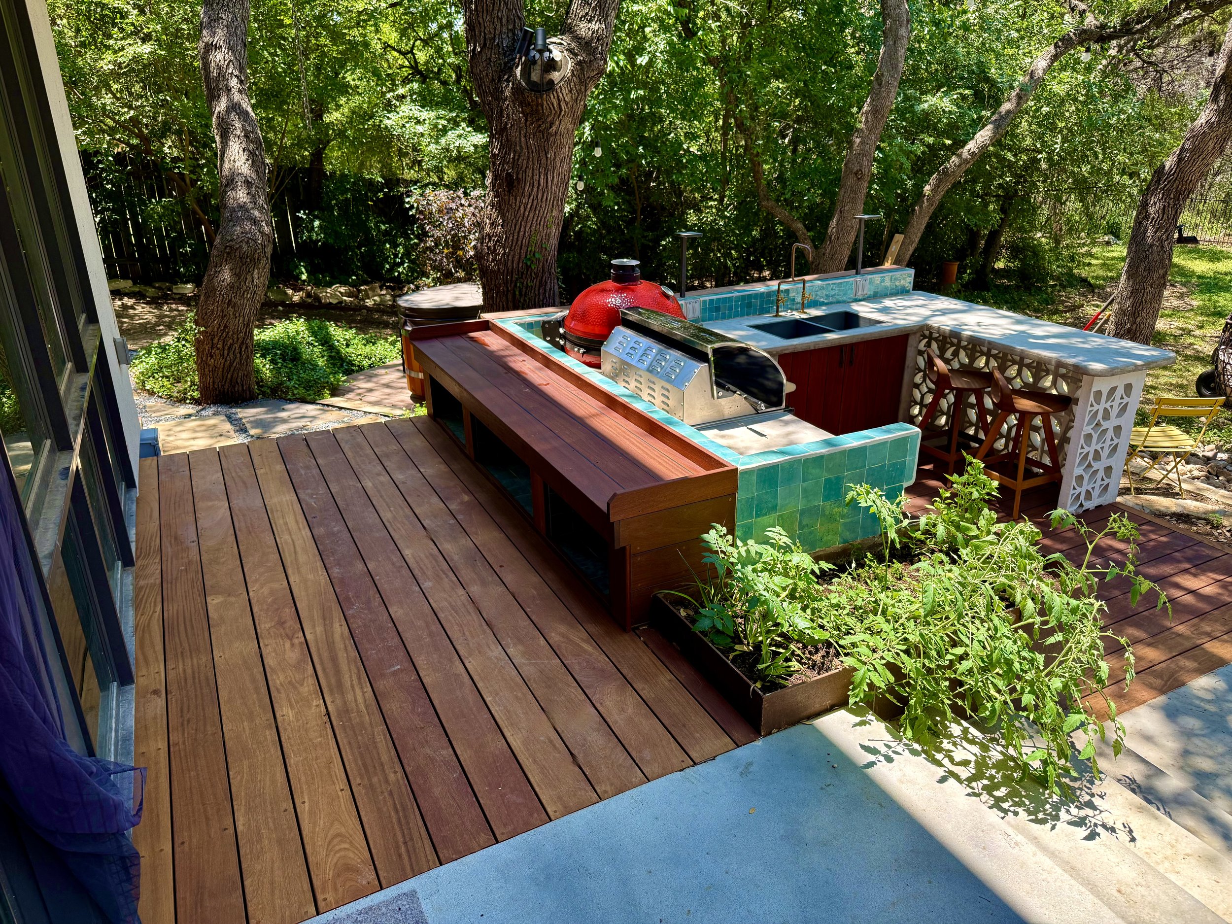 Outdoor patio area with a built-in outdoor kitchen featuring green tiles, a red kettle grill, and a bar with two chairs, surrounded by trees and plants.