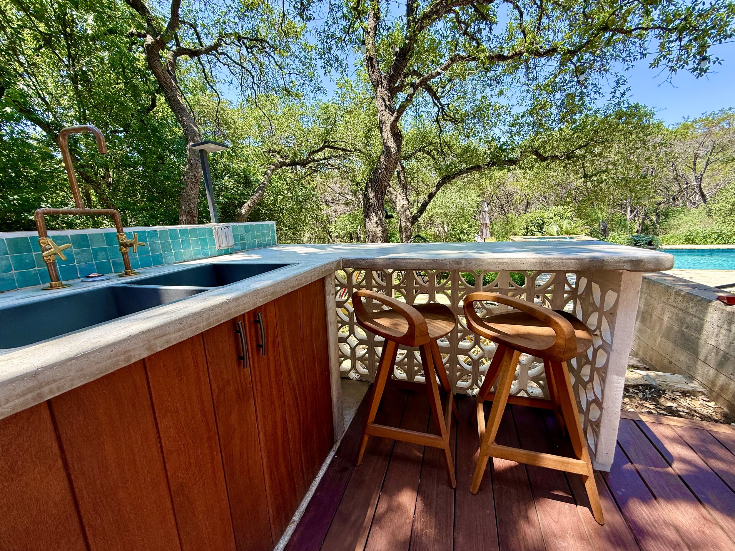Outdoor kitchen with a double sink, green tiles, wooden cabinets, two wooden bar stools, a white lattice wall, trees in the background, and a swimming pool to the right.