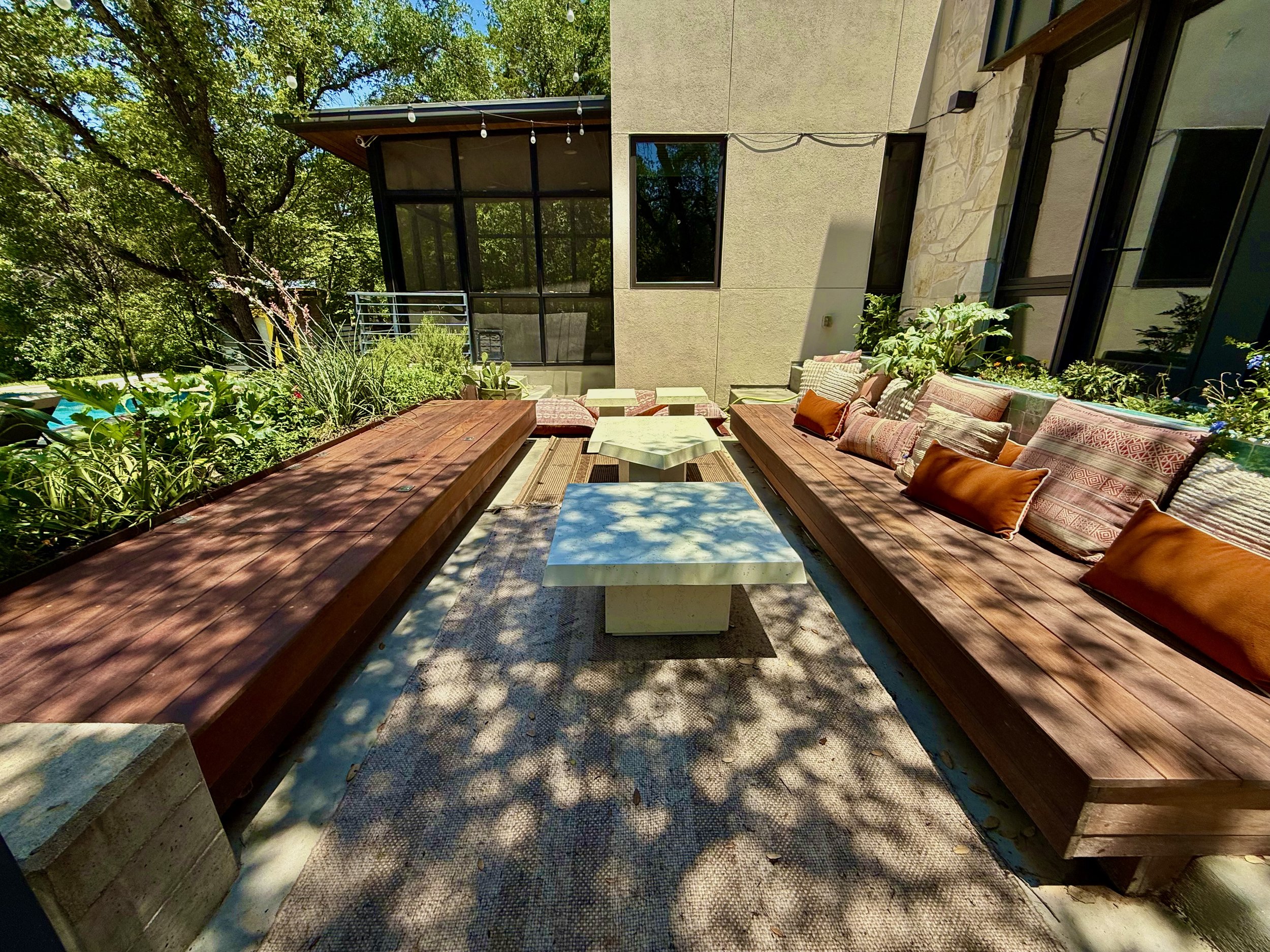 Outdoor patio with built-in wooden benches, cushion pillows, marble tables, surrounding green plants, and trees with shadows