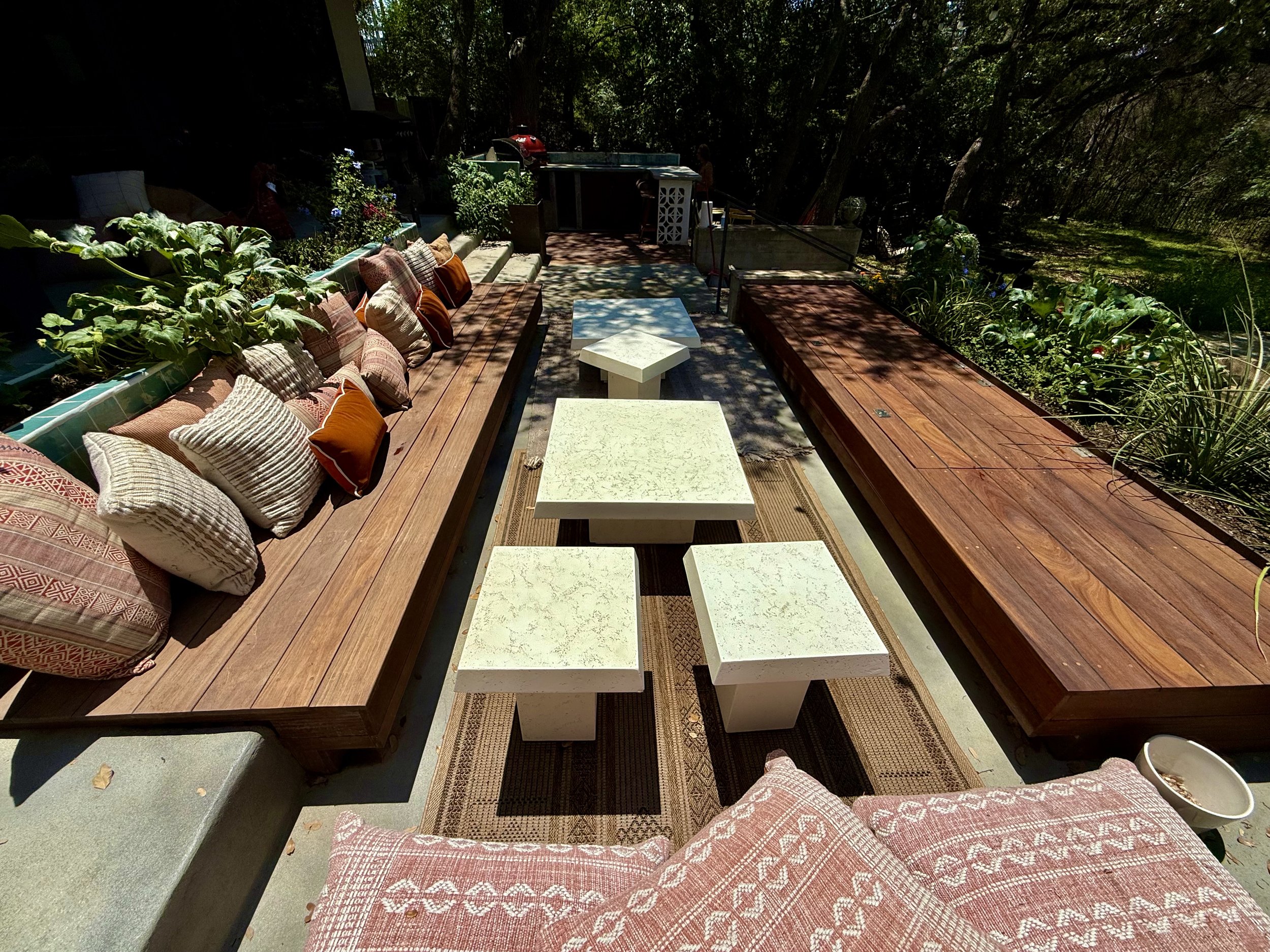 Outdoor seating area with wooden benches, white stone tables, cushions, and greenery in a shaded garden.