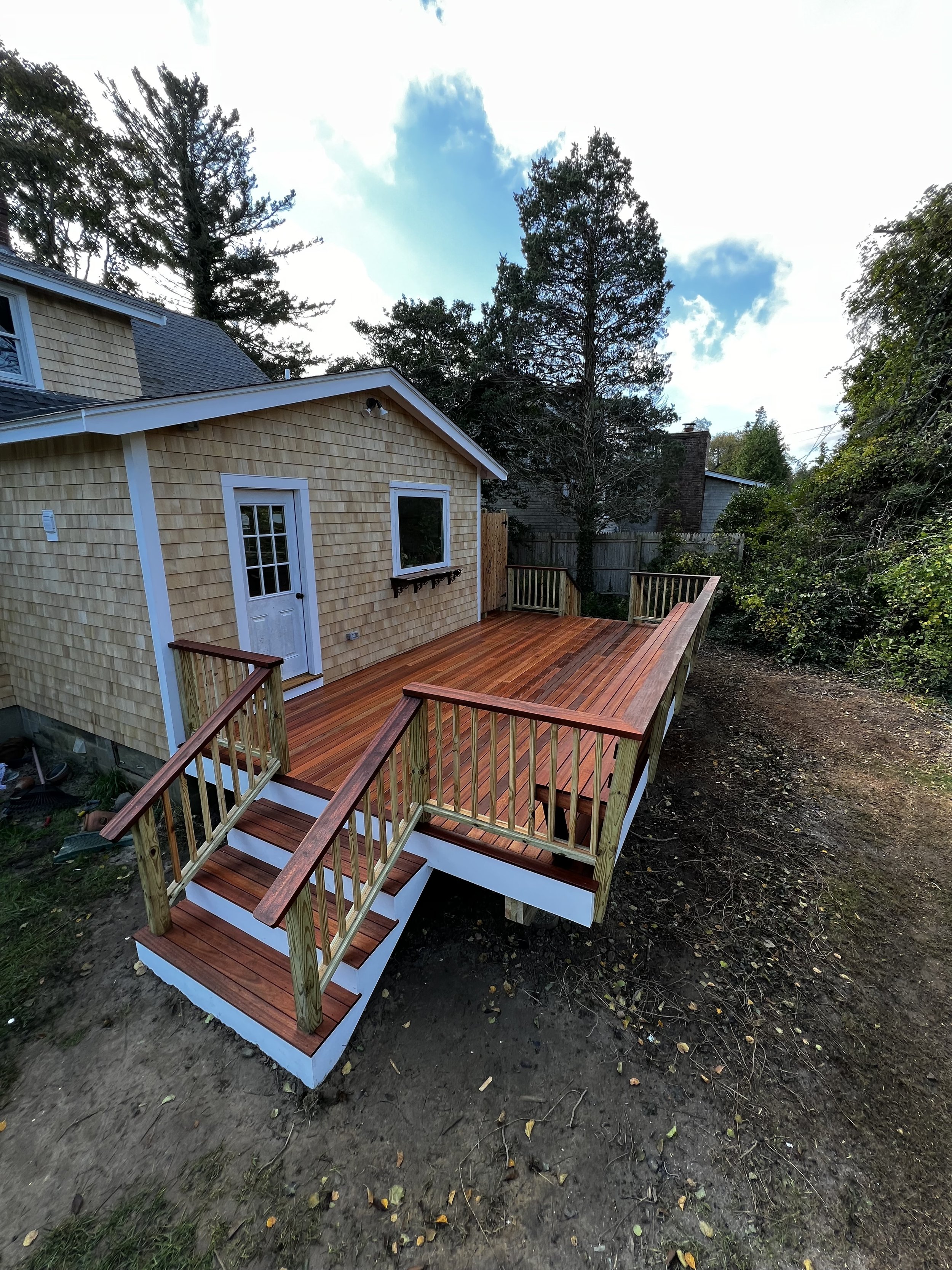 A newly built wooden deck attached to a house, with stairs leading down to a dirt yard, surrounded by trees and a wooden fence, under a partly cloudy sky
