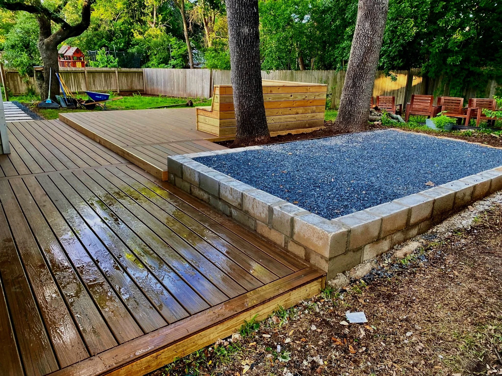 Composite ground level deck with a built-in cedar bench dining area and wrap-around stairs. A raised gravel pavilion was built beside the deck to accommodate a fire-pit and grill. A walkway leading off the property features custom concrete pavers.