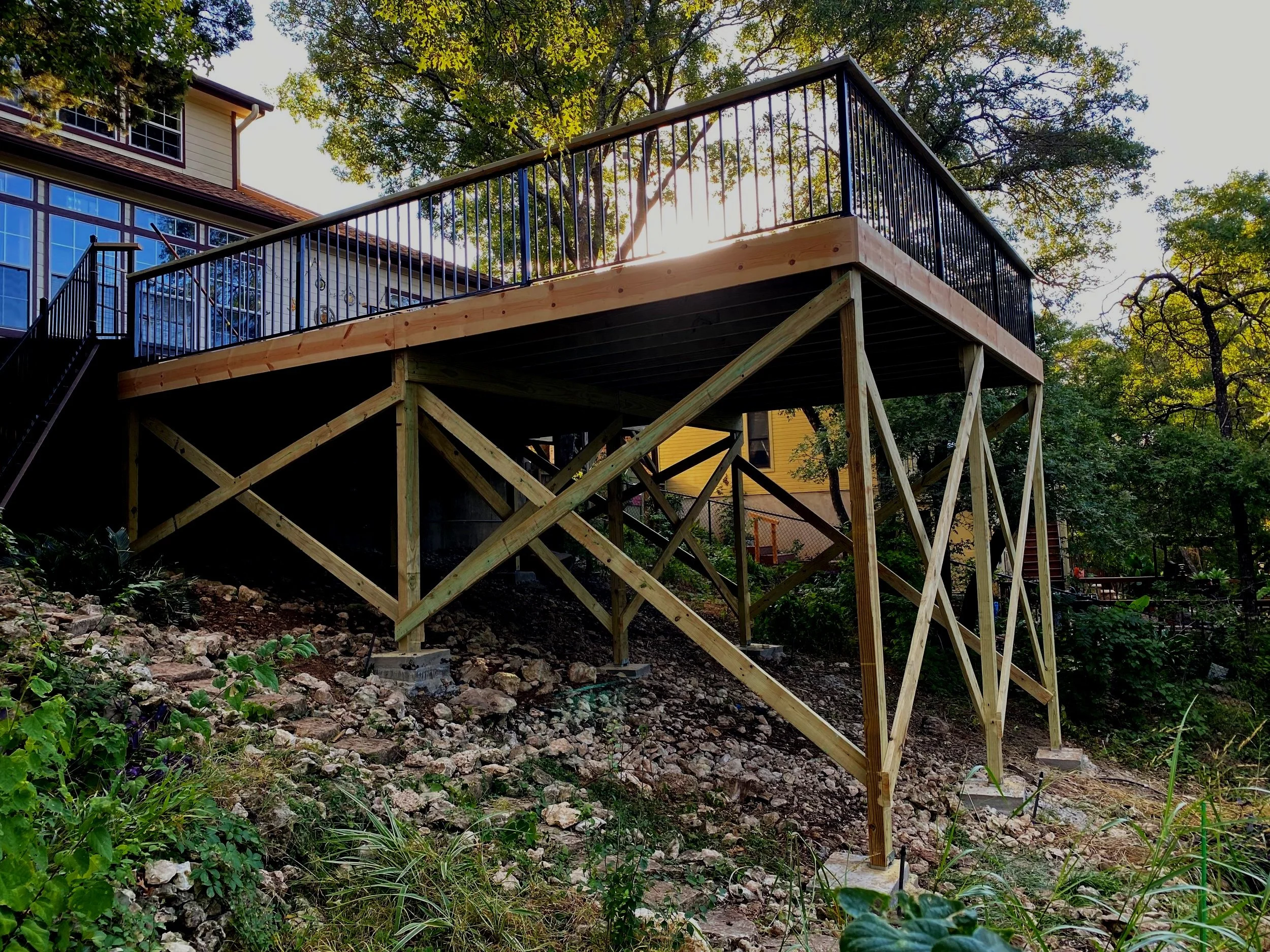 A newly built wooden deck with black metal railings elevated on wooden supports on a sloped yard, with trees and a house in the background.