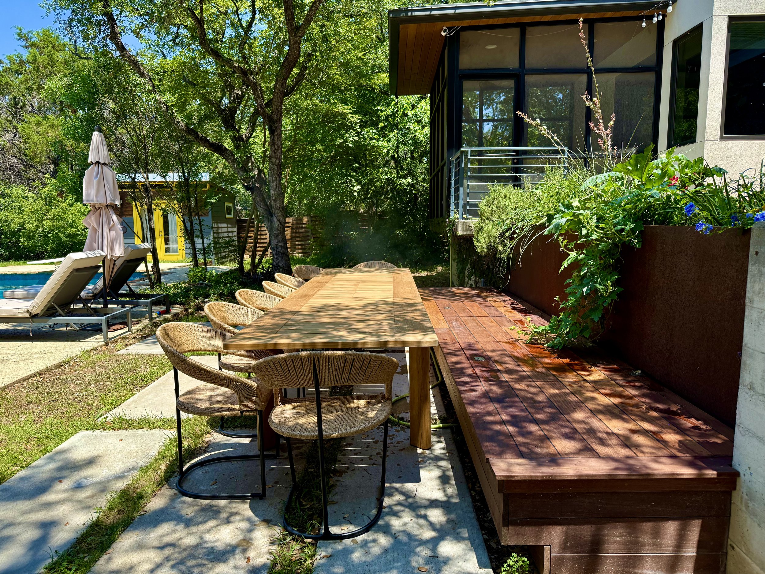 Outdoor backyard patio area with a large wooden dining table, wicker chairs, two lounge chairs near a swimming pool, lush green trees, potted plants, and a modern house with large windows and a balcony.