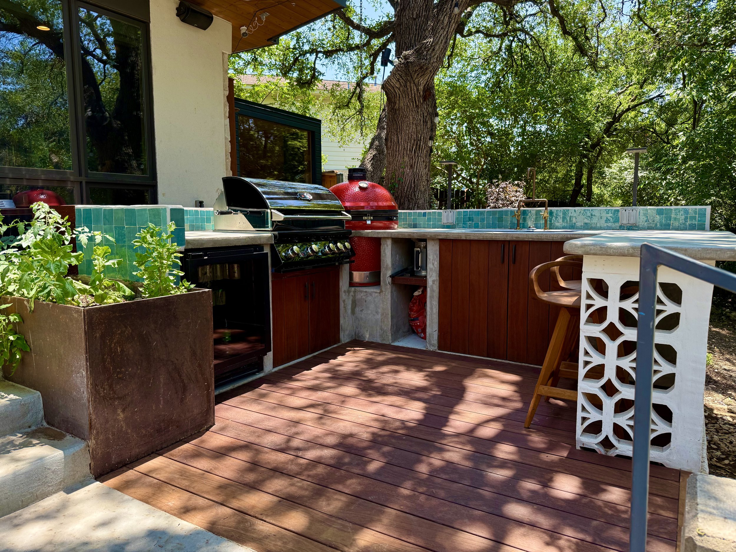 Outdoor kitchen with a grill, a red Big Green Egg, a tiled counter with a sink, wooden cabinetry, a white decorative table, a barstool, and nearby potted plants under trees.
