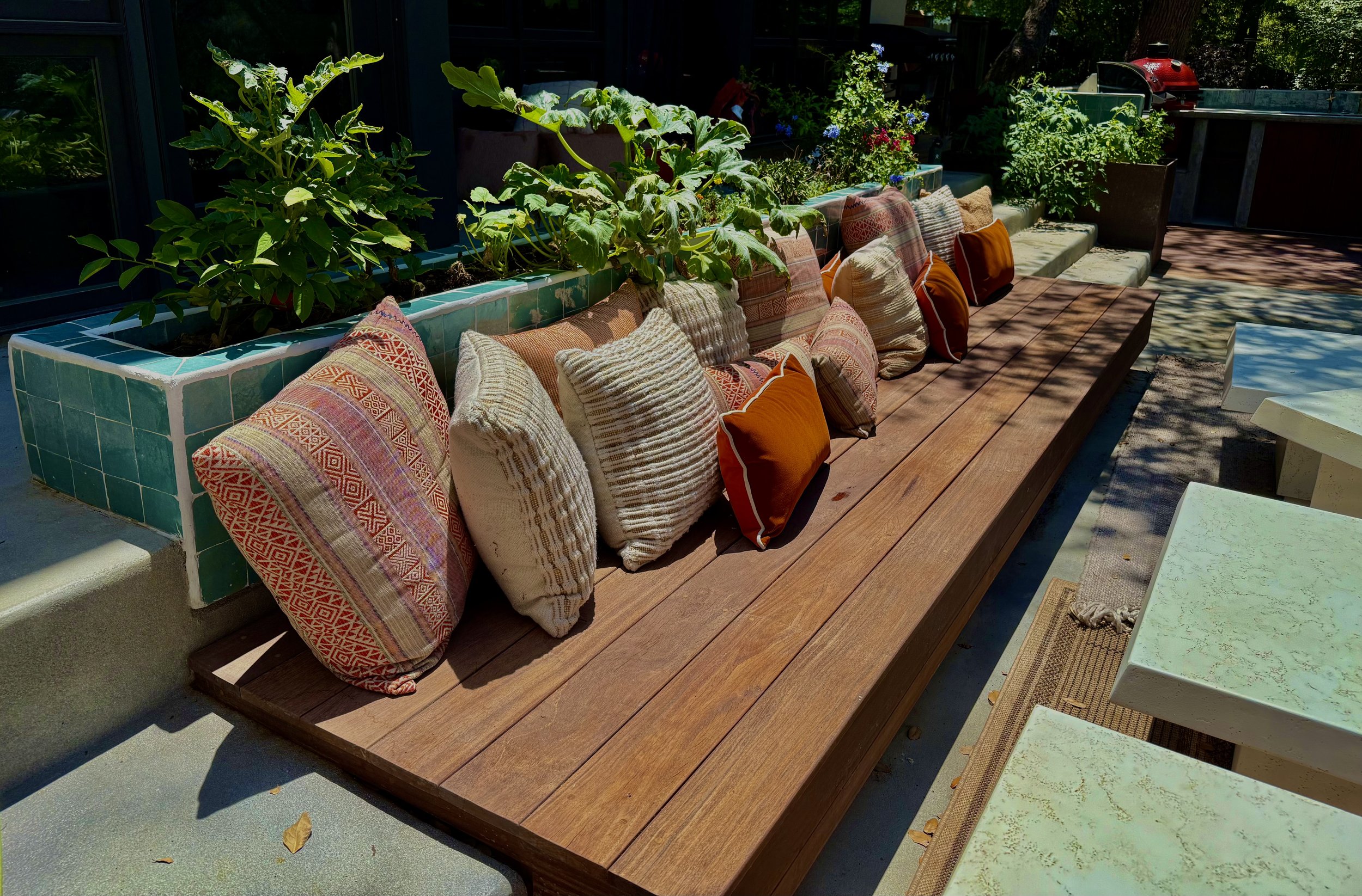 Outdoor wooden bench with colorful throw pillows, a green tiled planter box with plants behind it, and tables to the right, in a sunlit patio area.