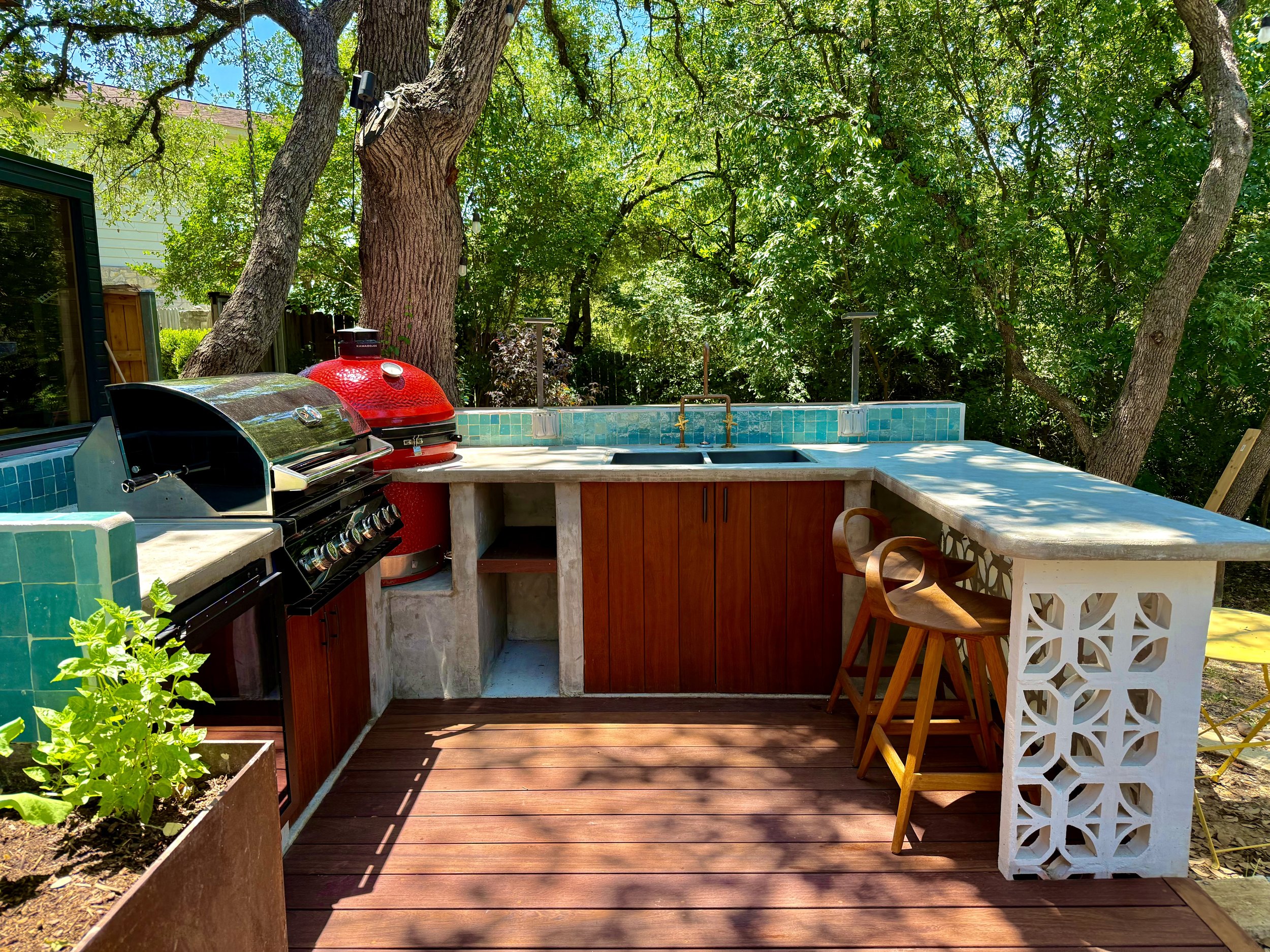 Outdoor kitchen with a built-in grill, red egg-shaped smoker, concrete countertops, wooden cabinet, two wooden bar stools, blue tile backsplash, surrounded by trees and greenery.