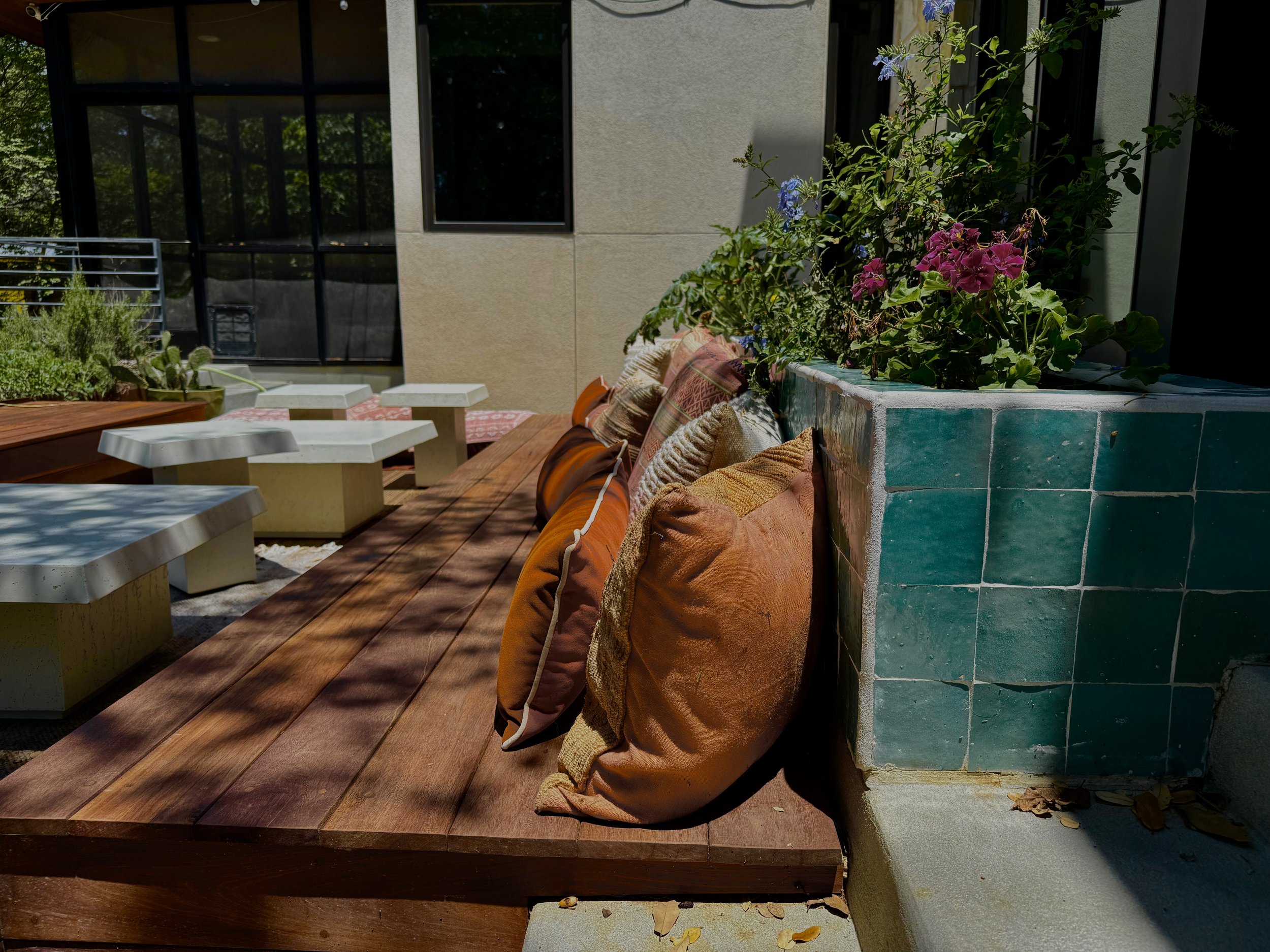 Outdoor patio with wooden decking, colorful pillows resting against a tiled planter with flowering plants, surrounded by modern outdoor furniture and a building with large windows.