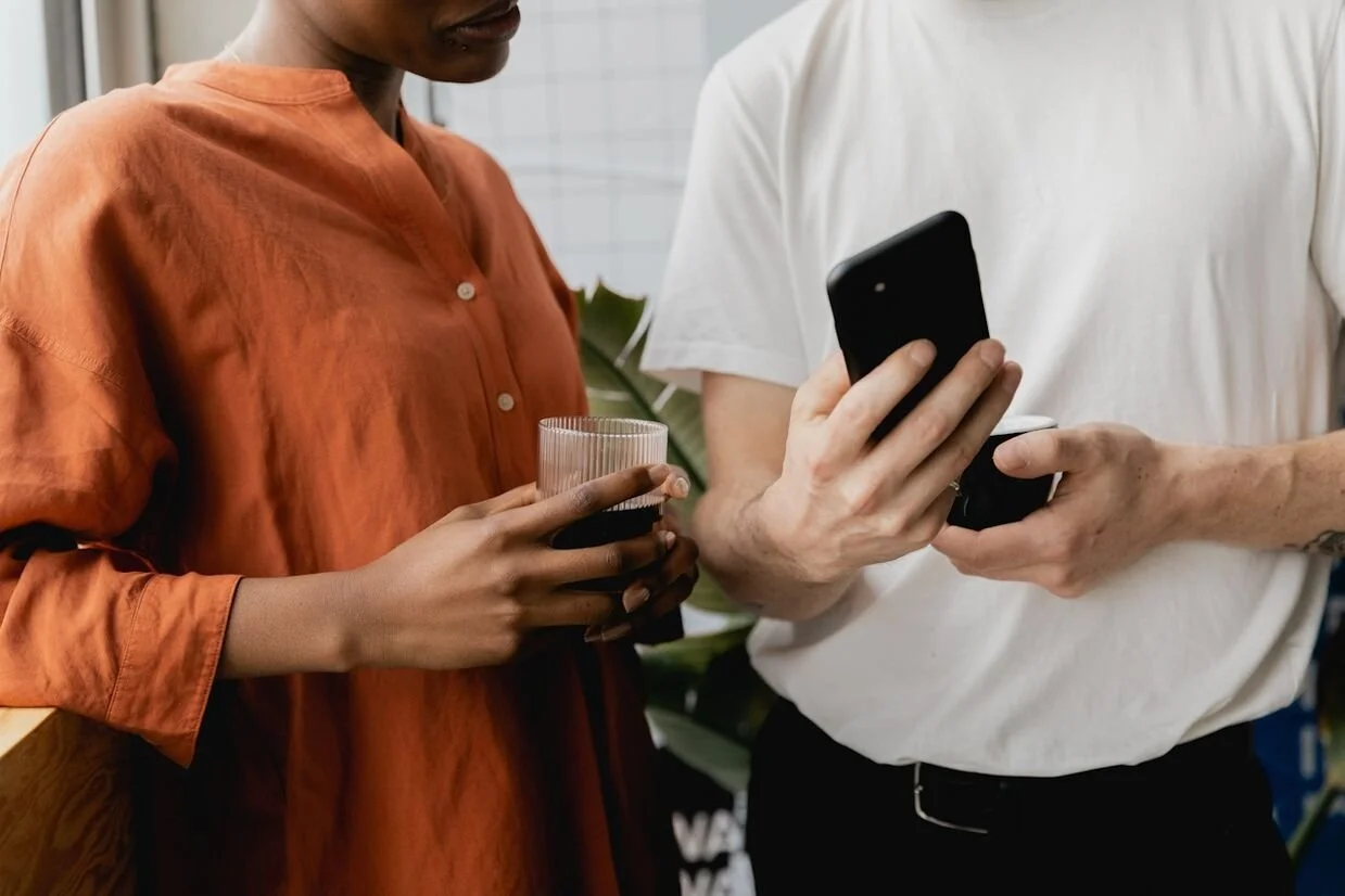 Two people engaged in conversation while one person is showing their smartphone screen to the other. Both have coffee cups in their hands.