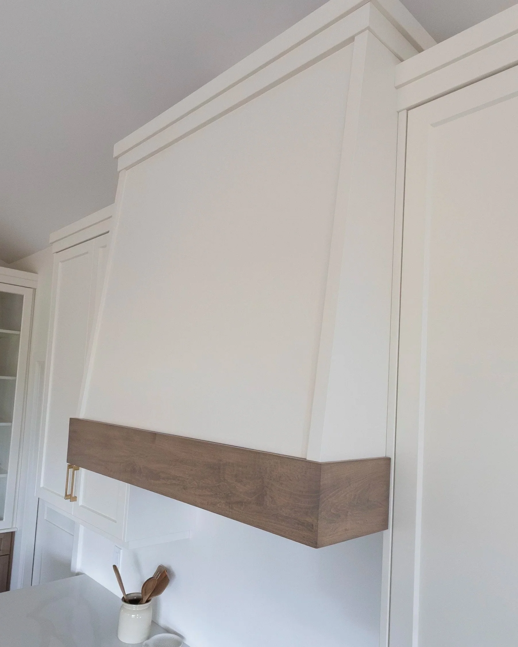 White kitchen or pantry area with a wooden floating shelf, white walls, and a partial view of a jar with wooden utensils, including a spoon and a spatula.