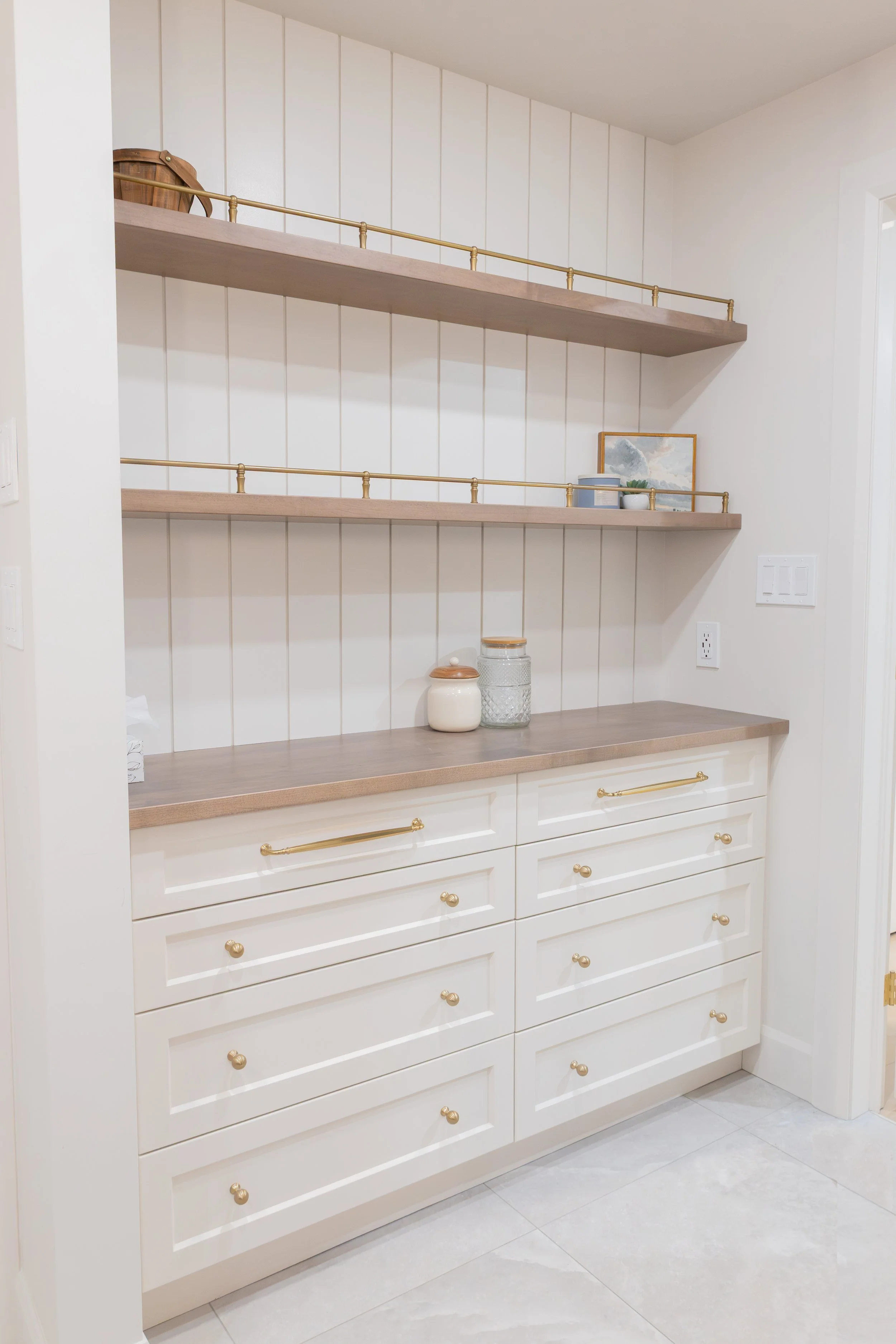 White built-in cabinet with gold handles, three upper open shelves with decorative items, and a lower cabinet with drawers in a bright room.