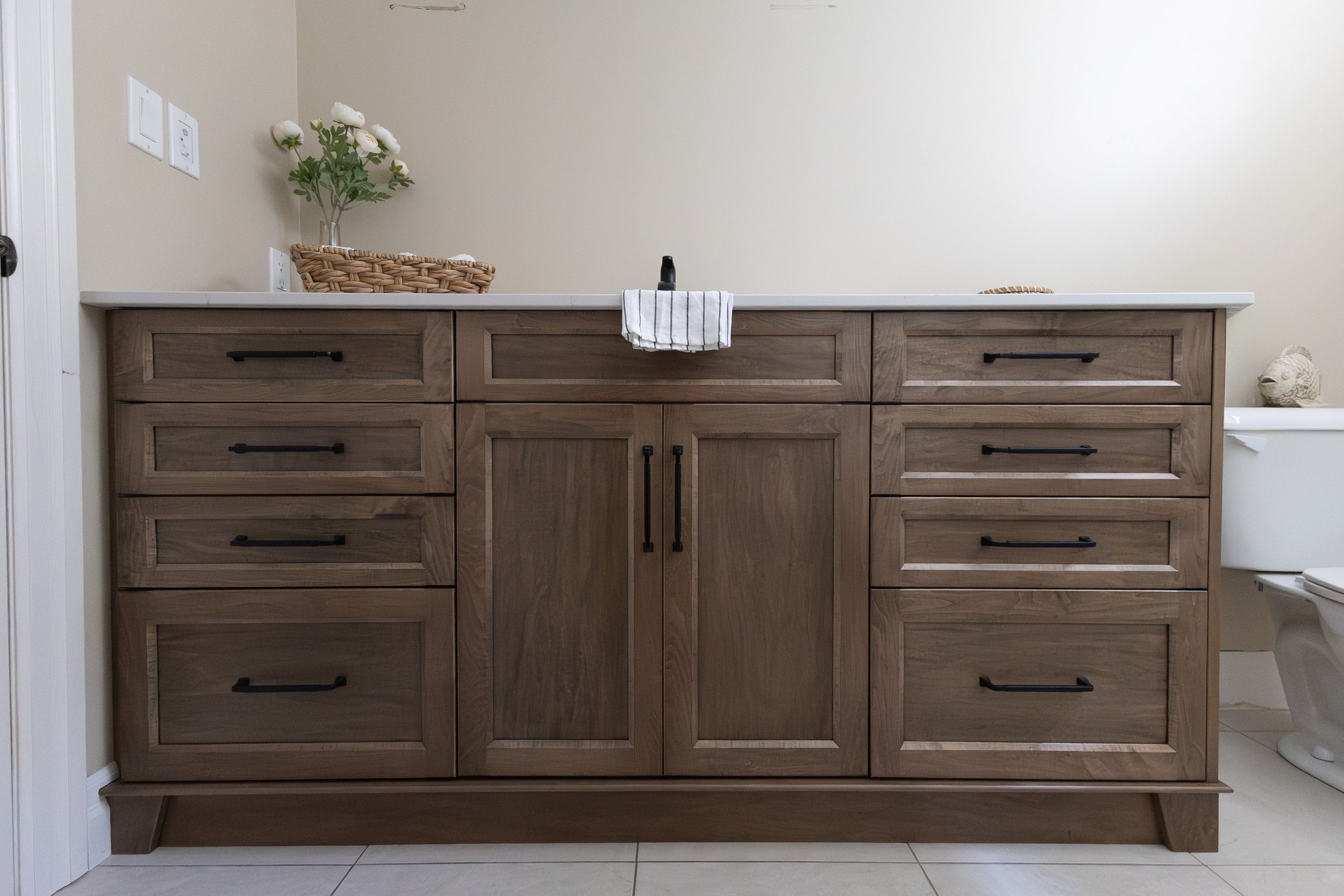 A wooden bathroom vanity with multiple drawers and cabinets, a white countertop, a small towel hanging over a black faucet, a basket with white flowers on the left, a small decorative animal on the right, and a partially visible toilet to the far rig