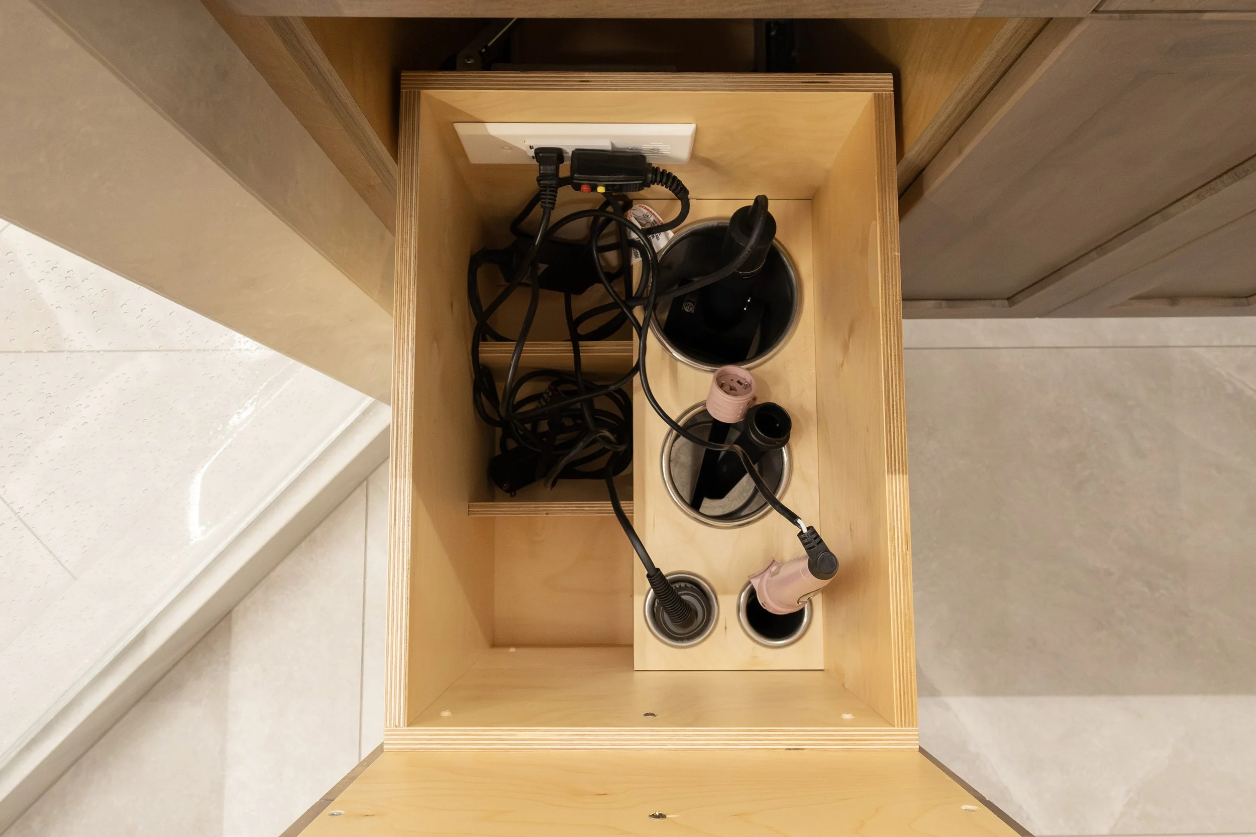 Empty wooden desk with cutouts for cables, showing scattered electrical cords, a power strip, and a hairdryer, with a wall socket visible on the wall.