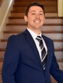 A man in a blue suit and striped tie smiling, standing in front of a wooden staircase.