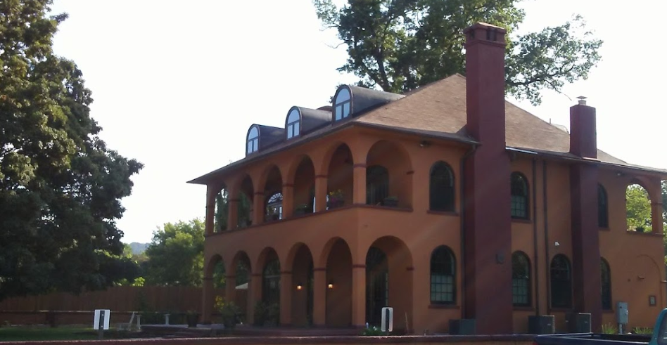 A large multi-story house with orange stucco walls, arched windows, and a brown roof is surrounded by trees and a wooden fence.
