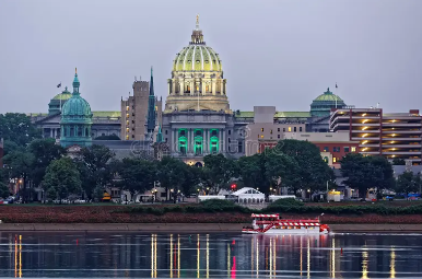 View of a historic building with a large green-domed roof, illuminated at dusk, reflected in a river with a boat passing by.