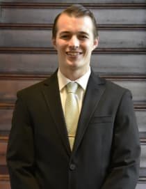 A young man in a black suit, white shirt, and light-colored tie, smiling, standing in front of a wooden background.