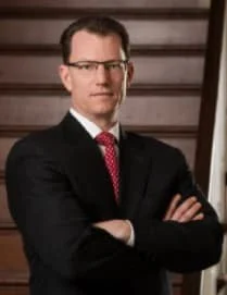 Man in black suit and pink tie standing with arms crossed in front of a staircase.