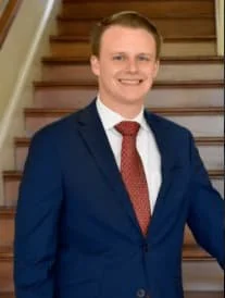Young man in a navy suit, white shirt, and red tie, standing in front of a staircase and smiling.