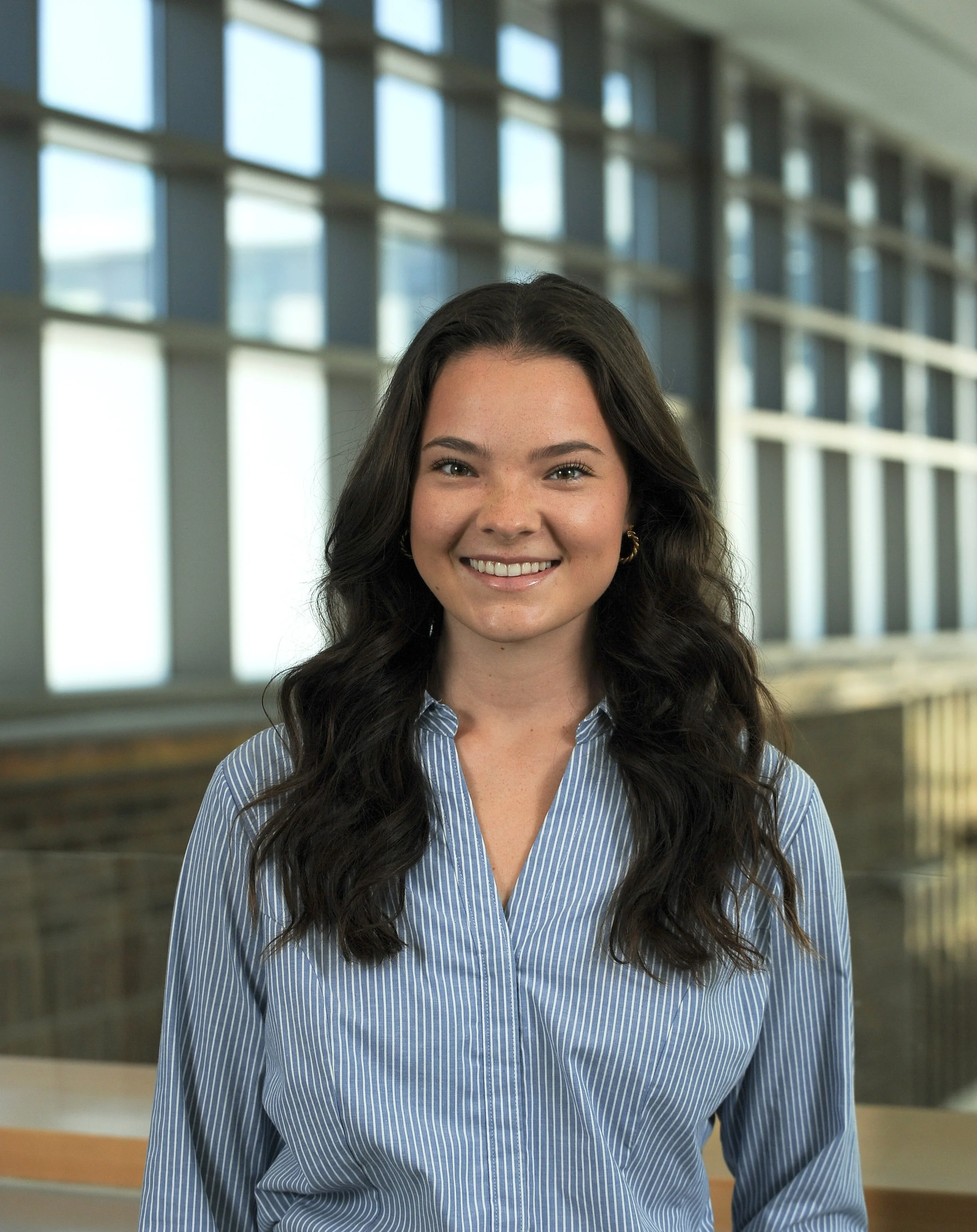 A smiling woman with long dark hair and earrings, wearing a blue and white striped shirt, standing indoors with large windows and a modern building interior in the background.