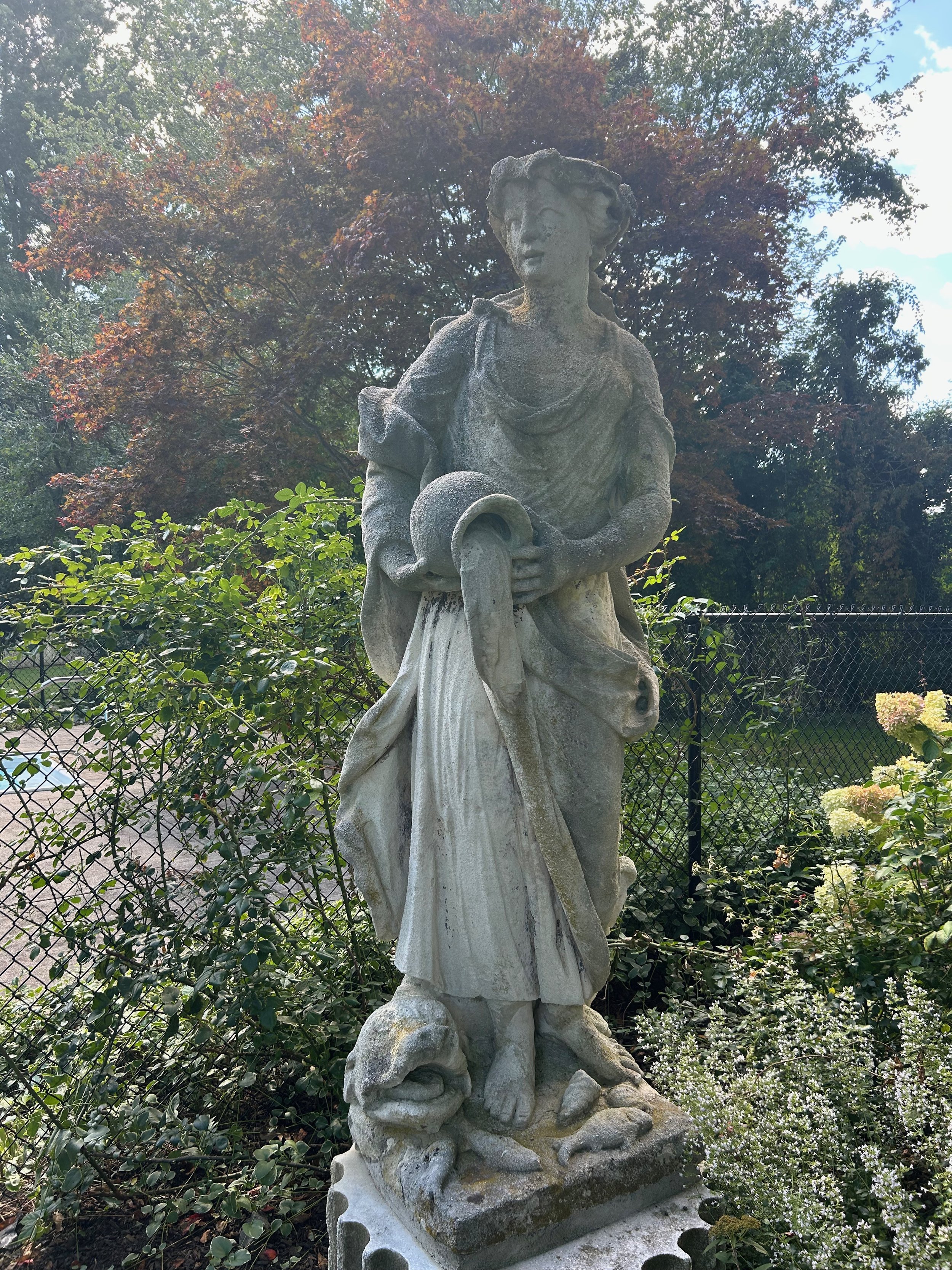 A stone statue of a woman in a flowing dress holding a large horn, set outdoors with trees and bushes in the background.