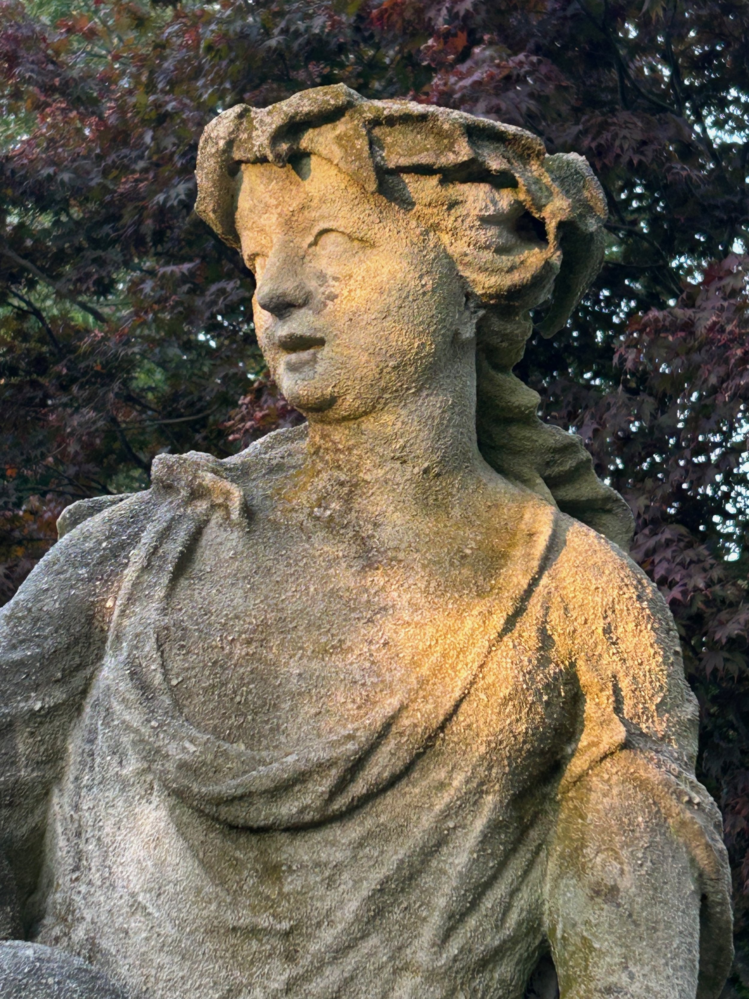 A weathered stone sculpture of a woman with flowing hair, set outdoors against dark foliage.