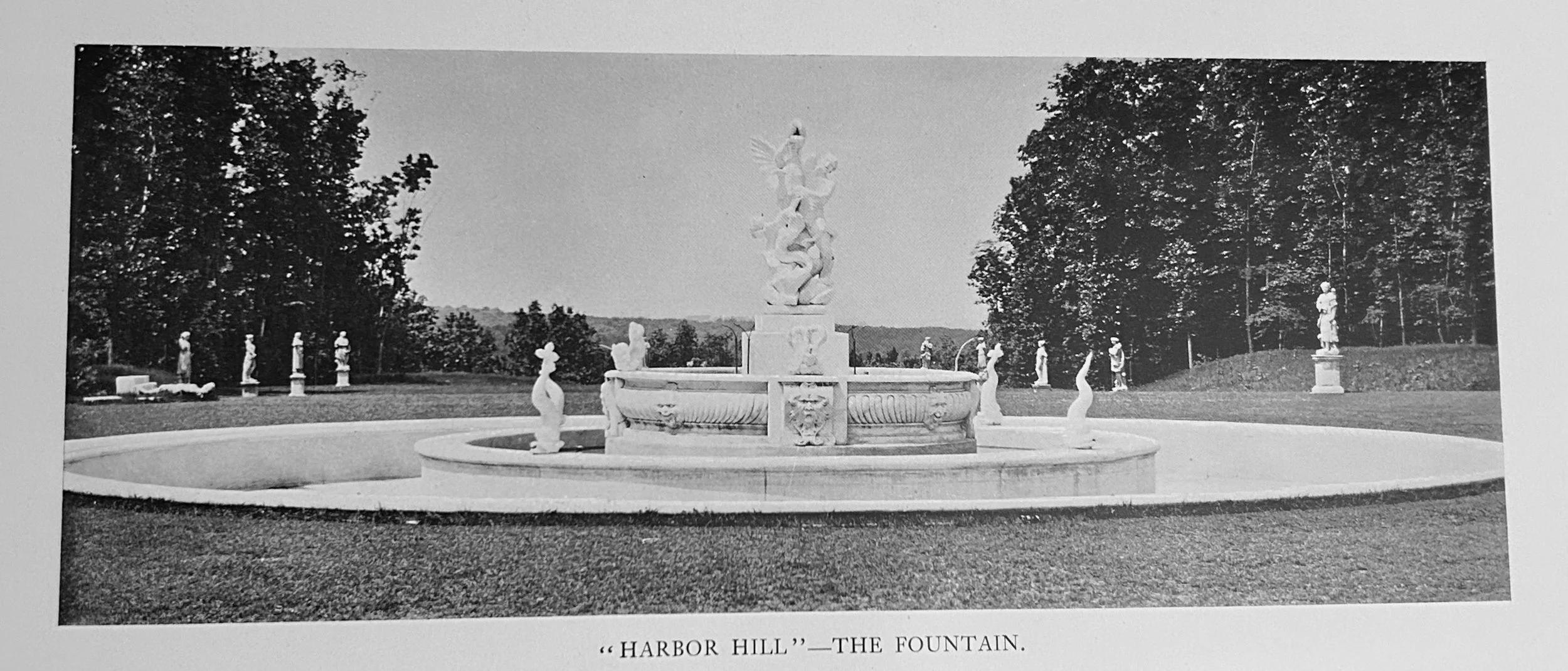 Black and white photograph of a decorative fountain titled 'Harbor Hill - The Fountain'. The fountain features a central sculptural group with figures and water spouts, surrounded by statues on pedestals in a park-like setting with trees in the backg