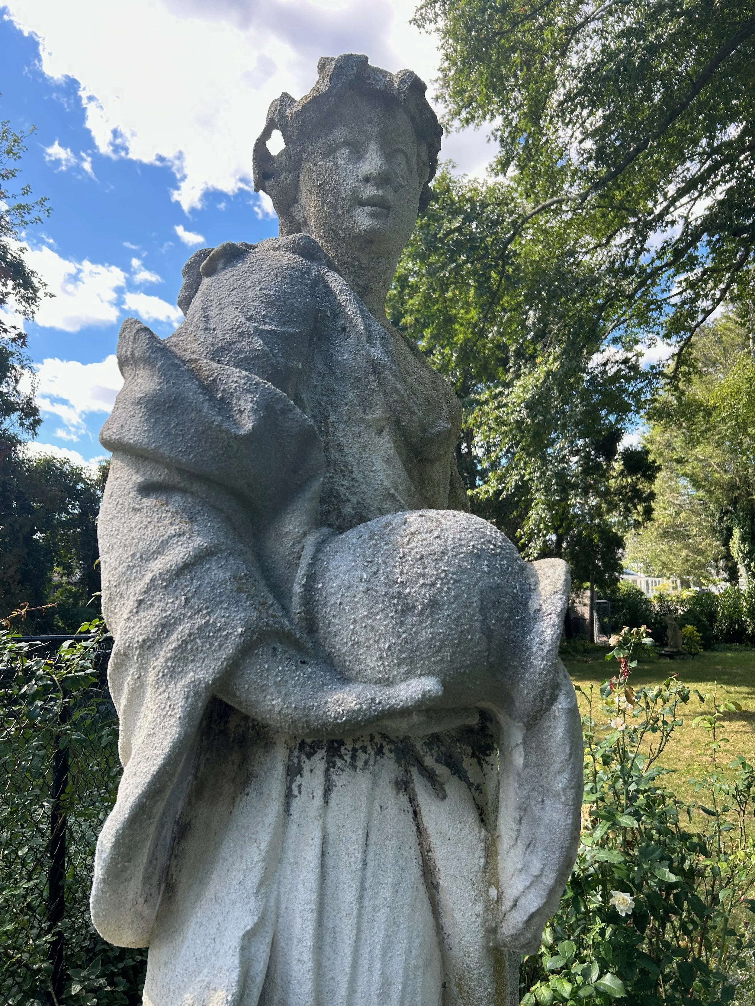 Close-up of a weathered stone statue of a woman with short hair, holding her hands near her chest, in a garden with green trees and a partly cloudy sky in the background.