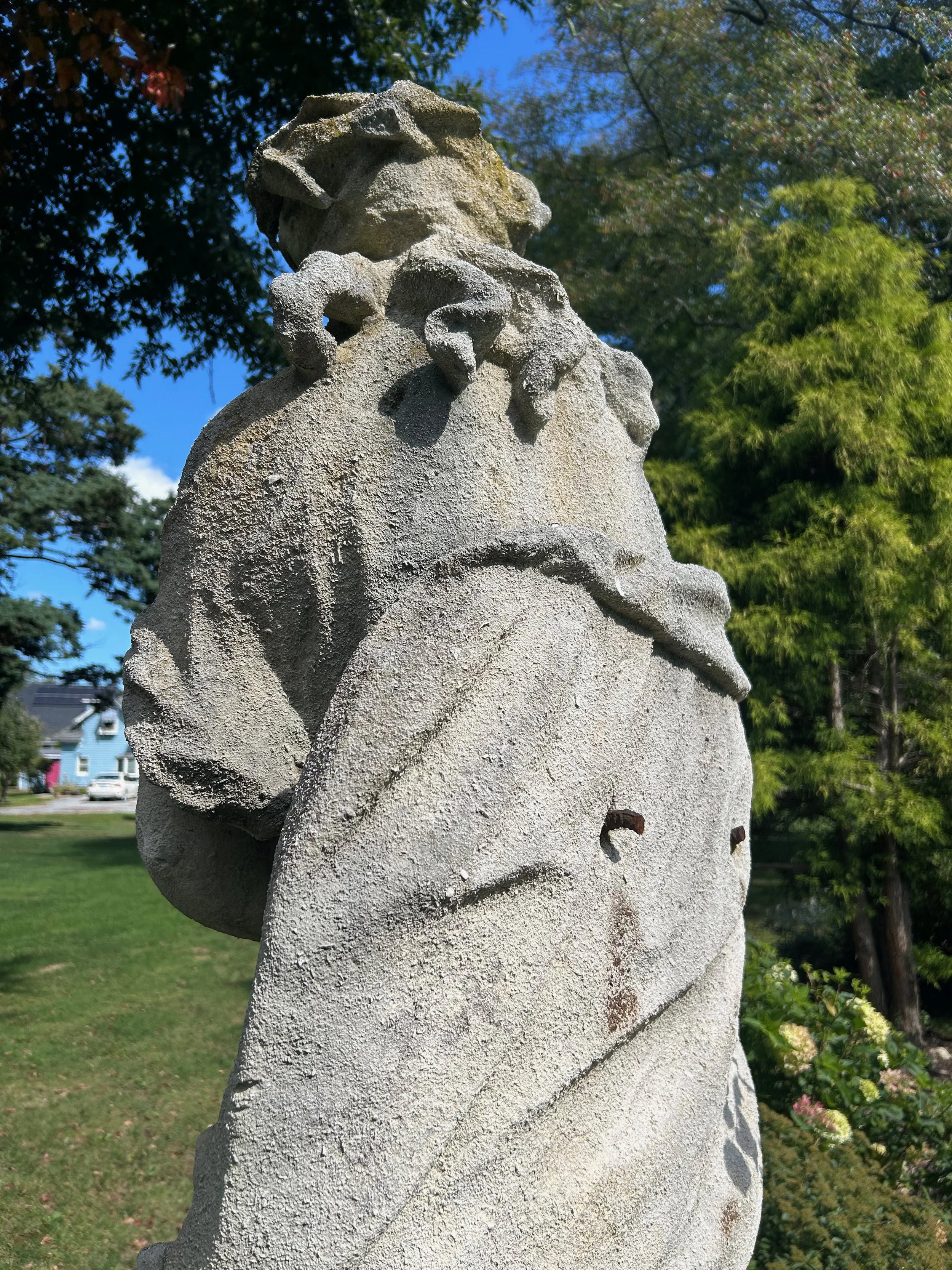Close-up of a weathered stone sculpture of a human face with a large beard, set outdoors with trees and houses in the background.