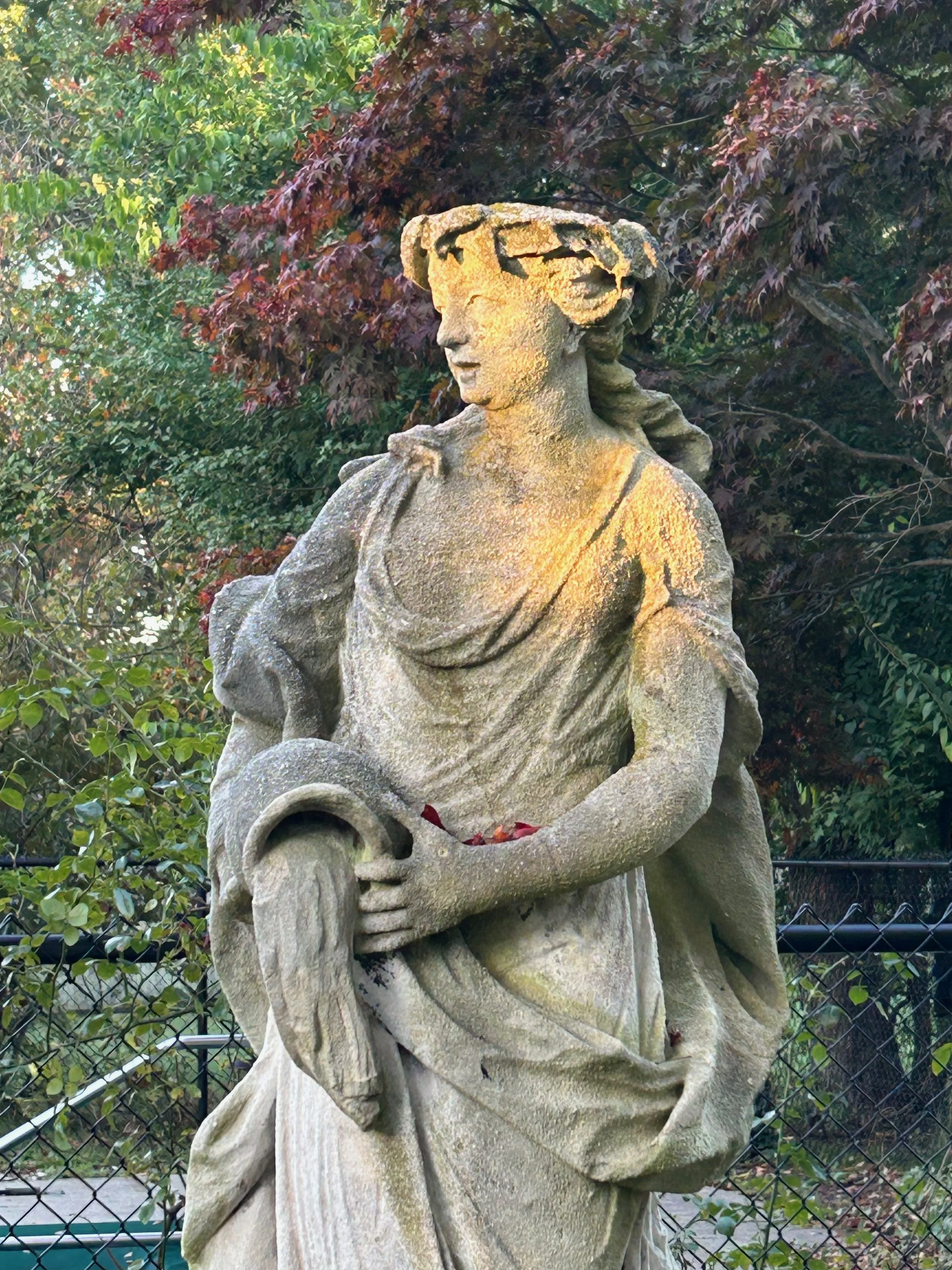 A stone sculpture of a woman, carrying a long folder with a handle, standing outdoors with greenery and trees in the background.