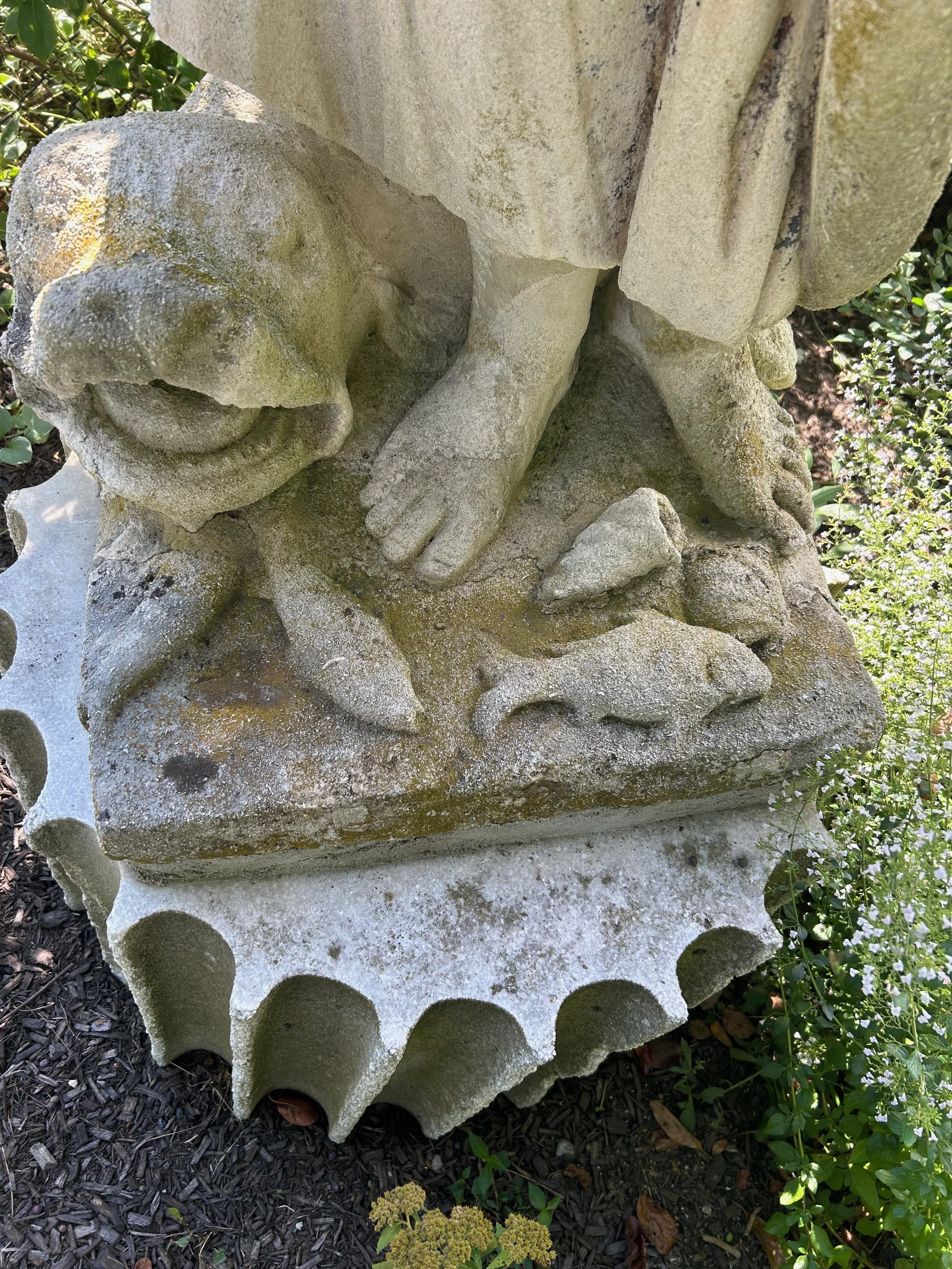 Close-up view of a weathered stone sculpture of a baby or angel lying down, with a detailed face, hands, and one leg visible, surrounded by greenery and small plants.