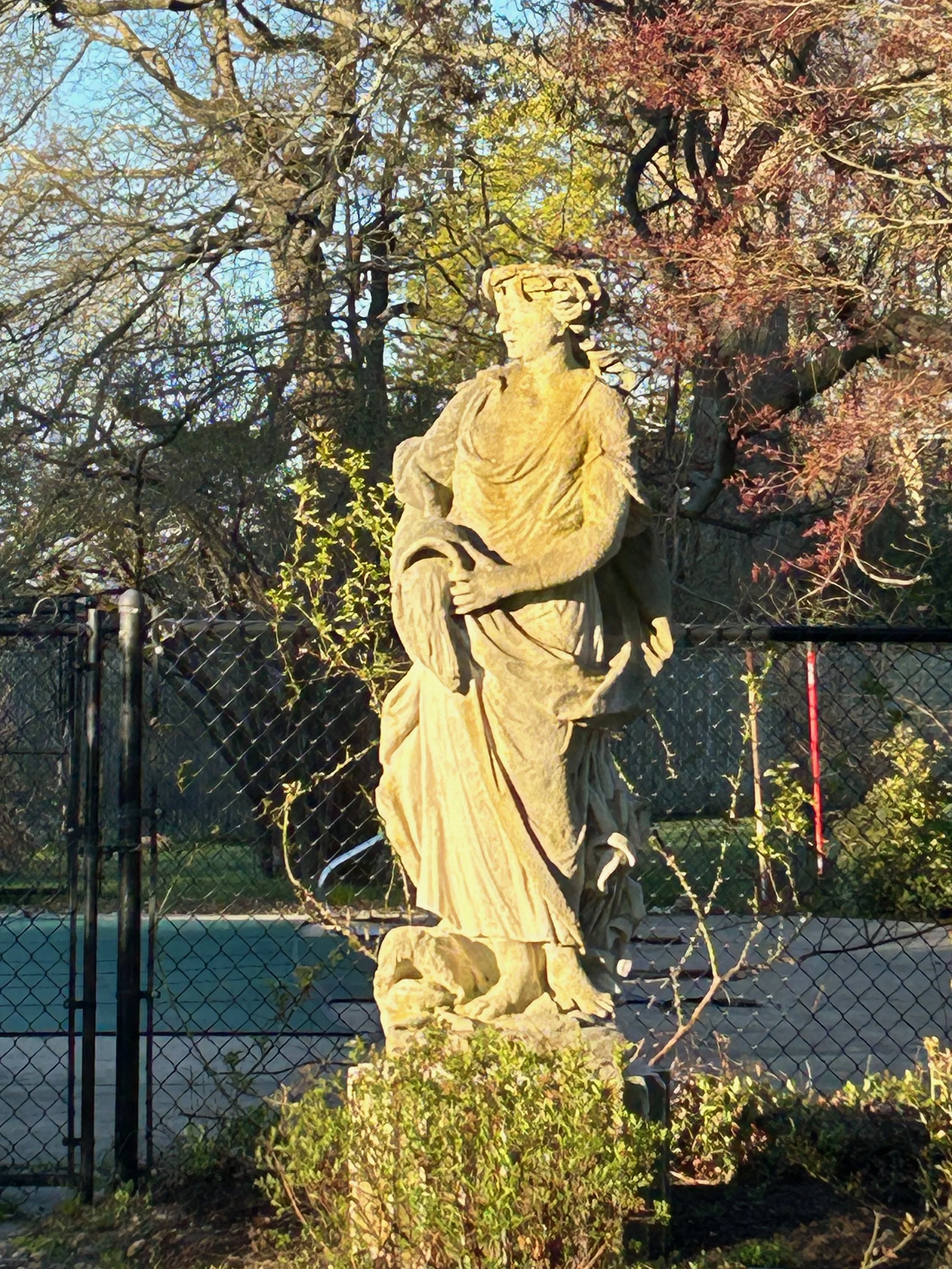 Statue of a woman with glasses and curly hair, standing outdoors in front of trees with autumn leaves, behind a black metal fence.