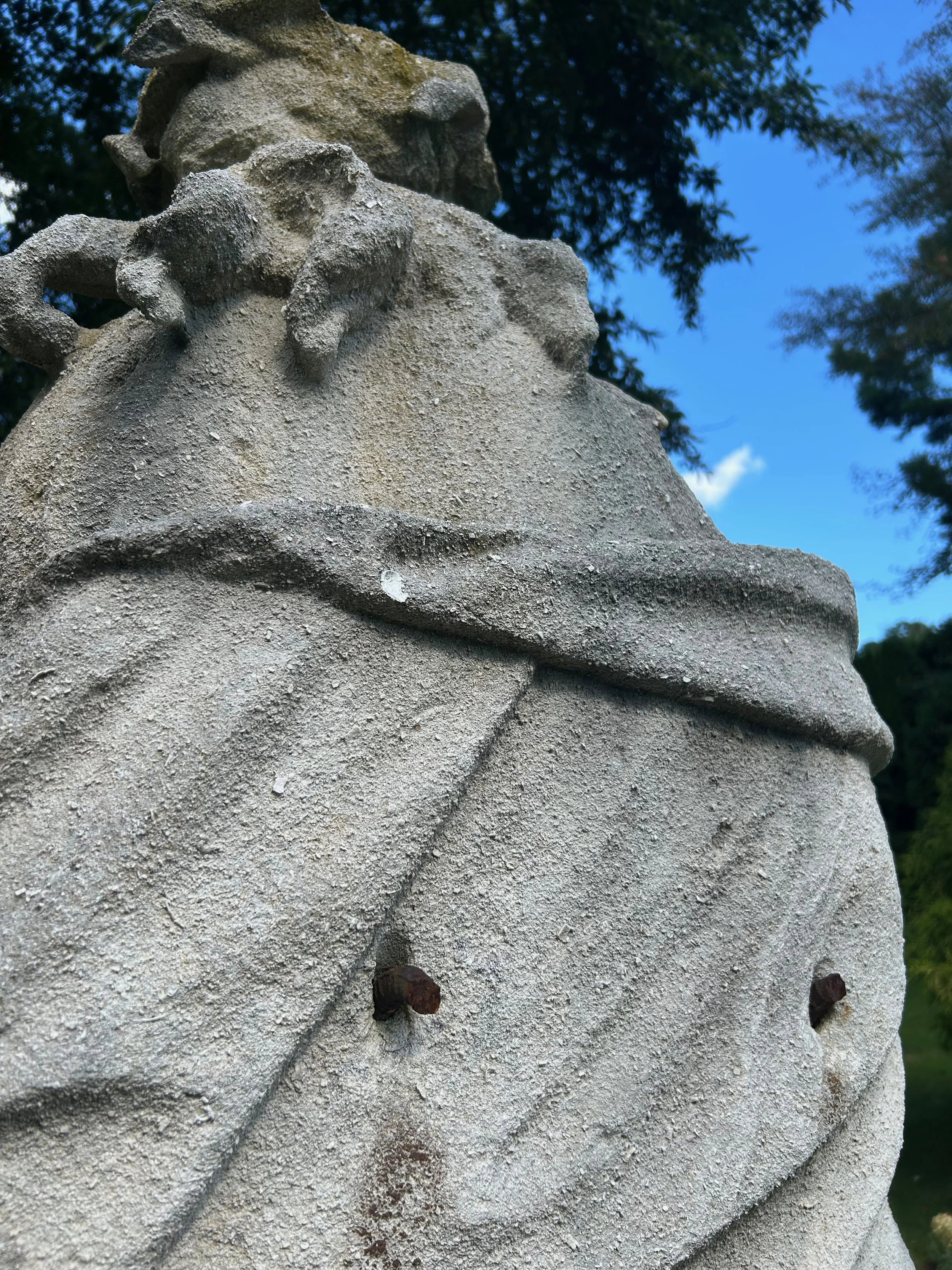 Close-up of a weathered stone sculpture of a face, with visible eye, nose, and textured surface, set outdoors against a background of trees and blue sky.