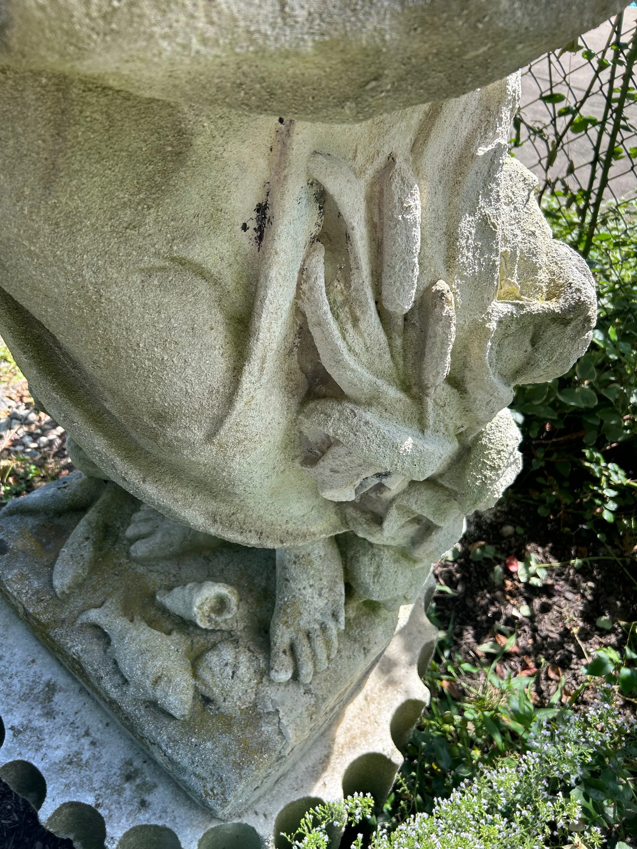 Close-up of a stone carved statue of a bird with intricate detailing, surrounded by green foliage and small flowering plants.
