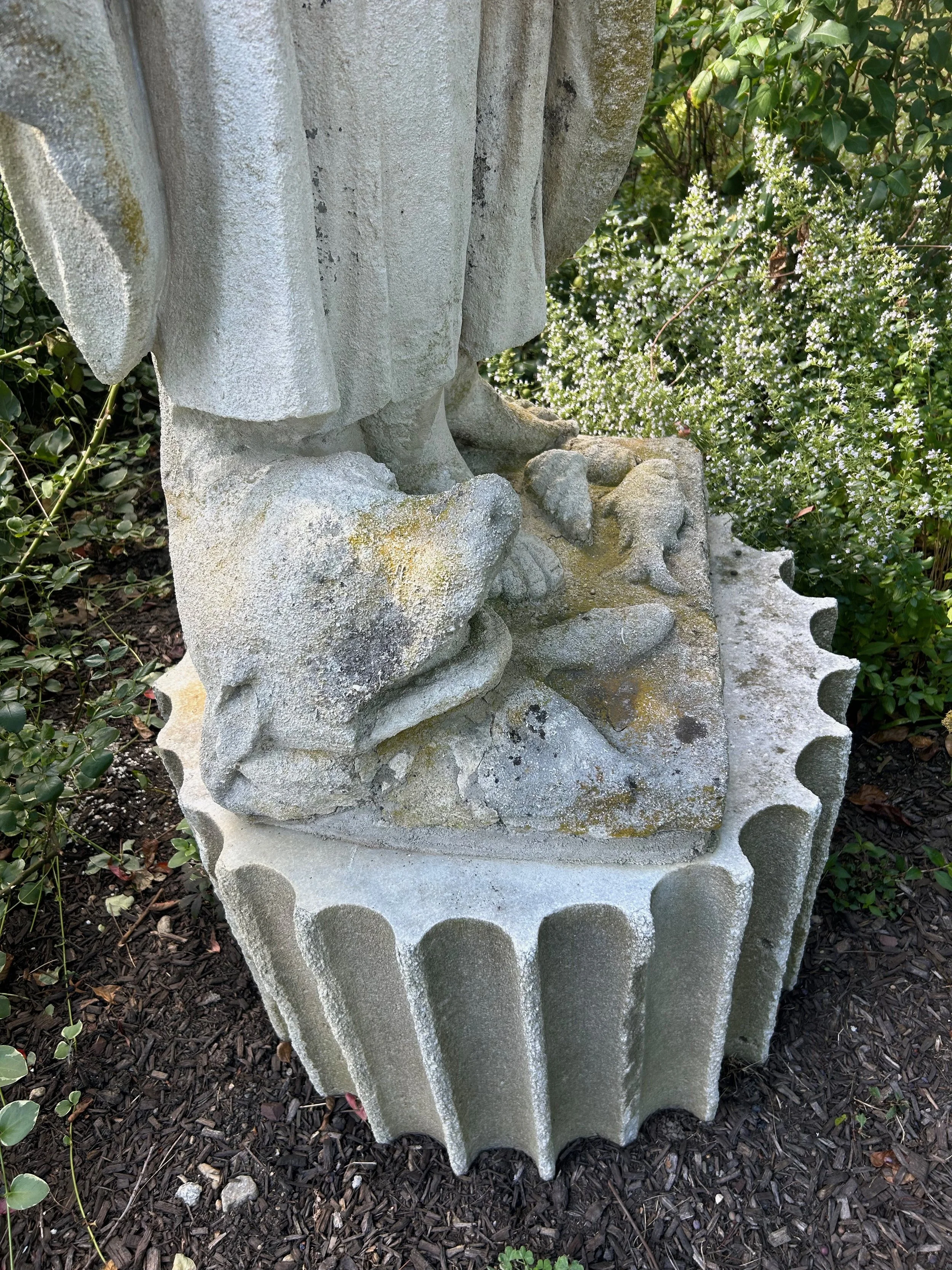 Close-up of a stone statue's lower part, showing legs and feet, surrounded by green bushes with small white flowers and dark mulch on the ground.