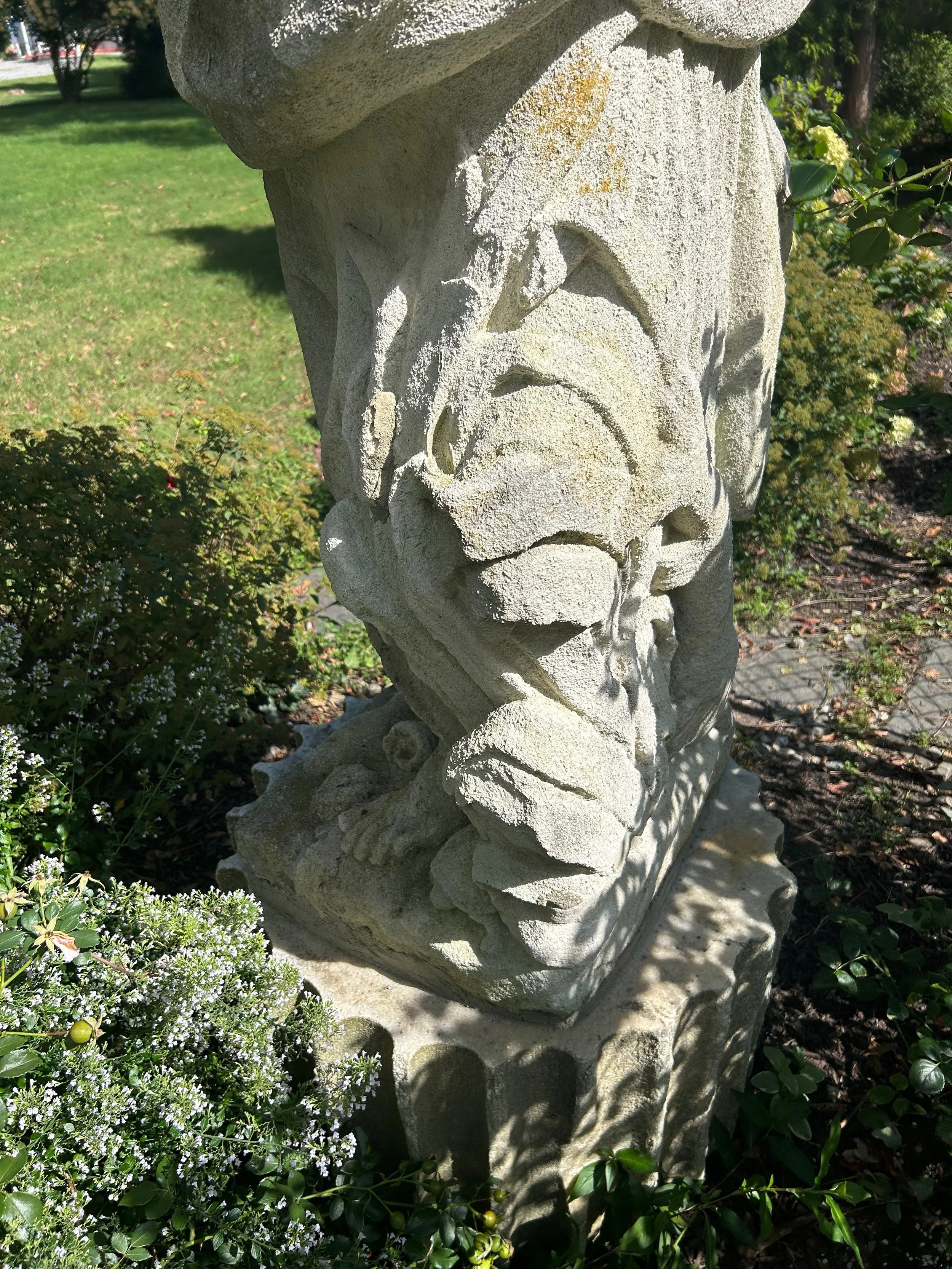 Close-up of a carved stone sculpture in a garden, surrounded by green plants and small white flowers, with trees and a lawn in the background.