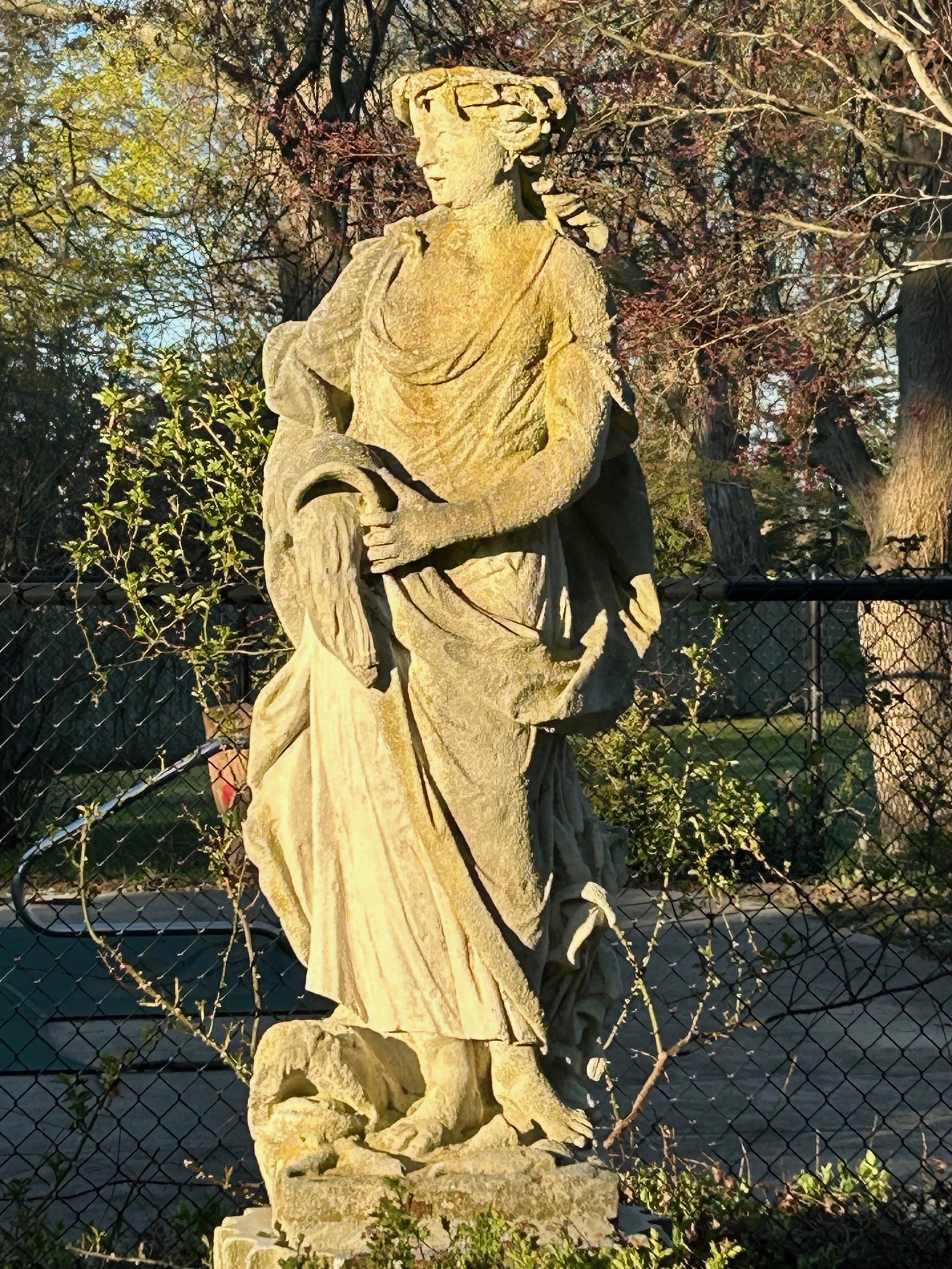 A stone statue of a woman with a hat, standing in front of trees with pink and green leaves, enclosed by a chain-link fence.