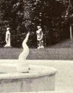 A white sculpture of a hand with fingers pointing upward, positioned in a fountain or circular basin, outdoors with trees in the background.