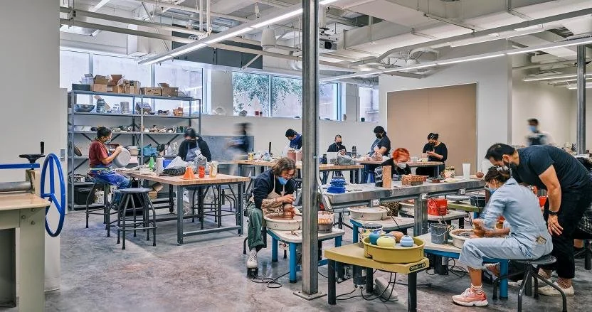 Ceramics classroom with multiple students using electric wheels to form clay vessels. Additional students work at metal workbenches around room.