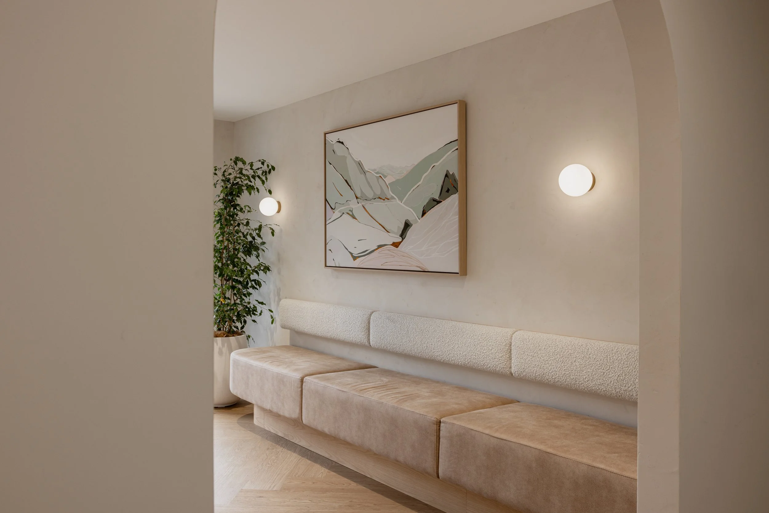 A minimalist waiting area with a beige cushioned bench, a large wall art piece depicting mountains, two round wall lights, and a potted plant in a white pot against a neutral wall.