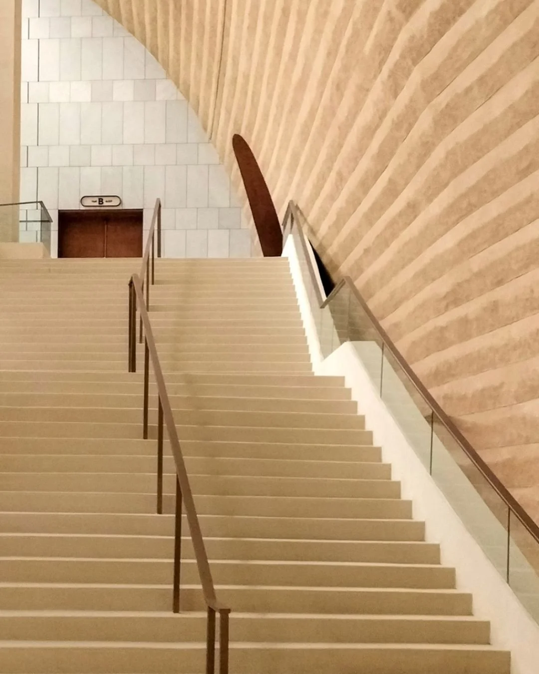 A staircase with beige steps and a glass railing, leading upwards. To the right, there is a wall with a wooden panel design, and at the top, there is a section of tiled wall with a closed brown door labeled 'B'.