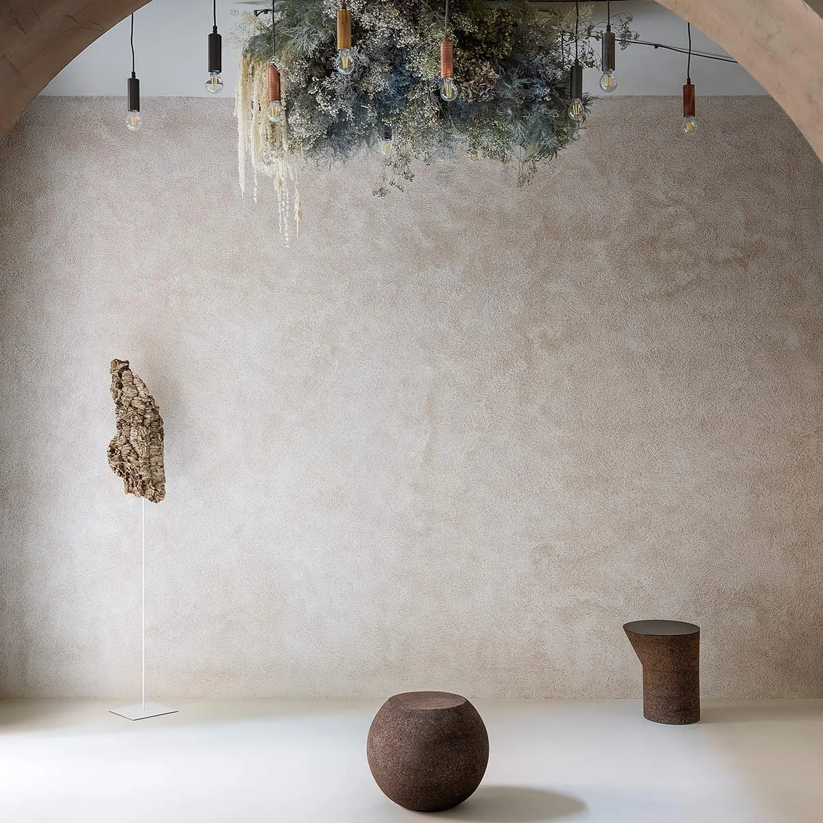 Minimalist interior with a textured beige wall, hanging light bulbs, a large greenery and floral installation on the ceiling, a sculpture on a stand, and two brown textured furniture pieces, one round and one cylindrical.