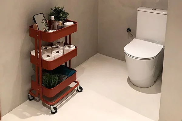 A small bathroom with a white toilet and a red rolling cart holding toiletries, a mirror, and decorative plants.