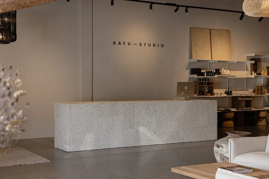 Interior of KAYU STUDIO with a minimalist aesthetic, featuring a textured white reception desk, a wall sign with the studio's name, a computer on the shelf, and modern decor elements.